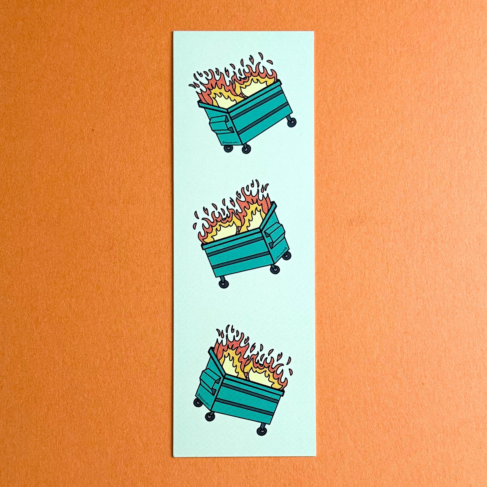 Front of Dumpster Fire bookmark- three hand drawn dumpster fires tossed and reflected on a light green background. The bookmark is on a solid orange background.