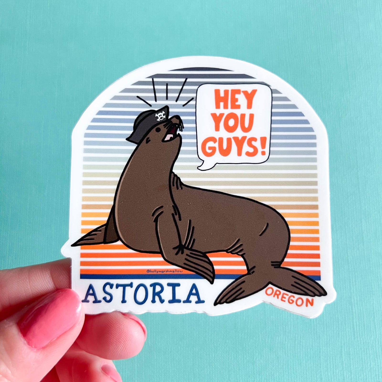 Sticker of a seal with text 'Hey You Guys! Astoria, Oregon' held against a light blue background.