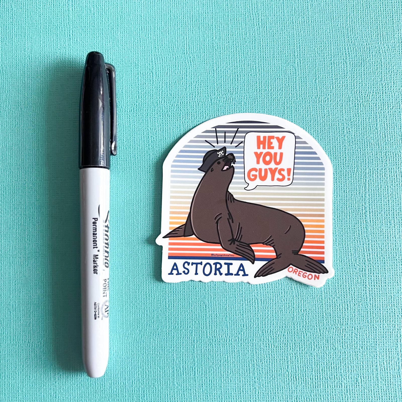 Sticker with a seal and text 'Hey You Guys! Astoria, Oregon' next to a Sharpie on a teal background