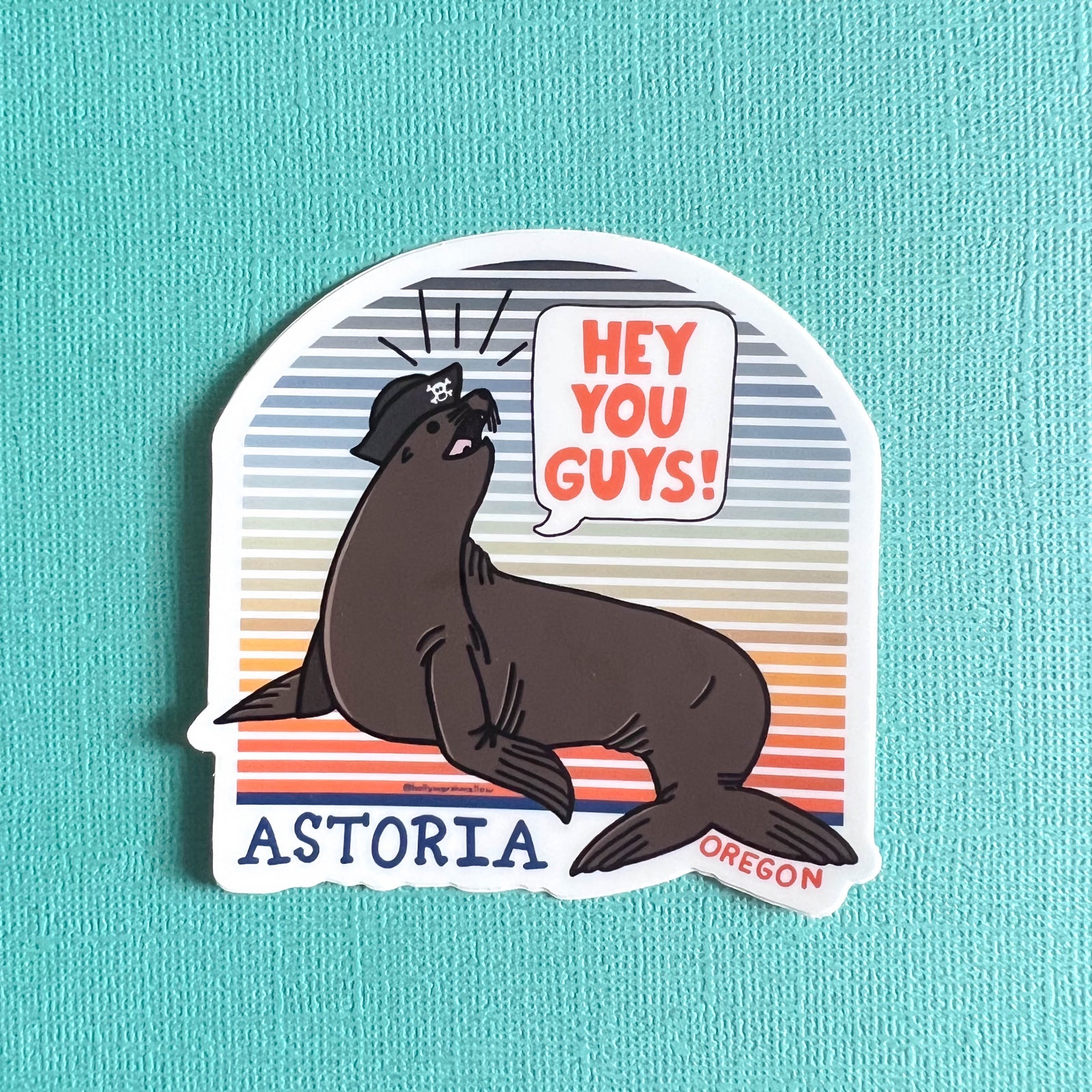 A magnet featuring a hand-drawn illustration of a sea lion wearing a tricorn hat with a skull, and the phrase 'HEY YOU GUYS!' above. The background of the sticker is striped with colors gradient, and the word 'ASTORIA' is written at the bottom.