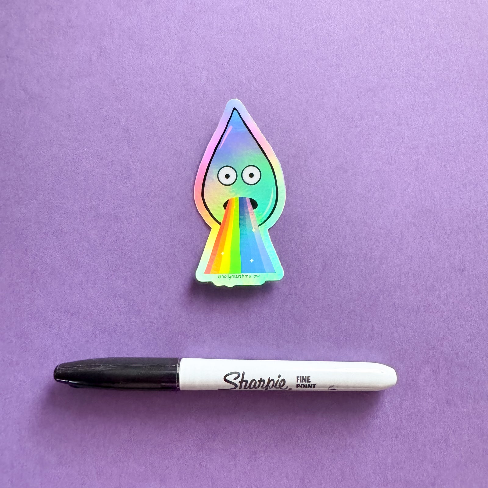 Holographic sticker of a raindrop with rainbow barf on a purple background, next to a Sharpie marker.