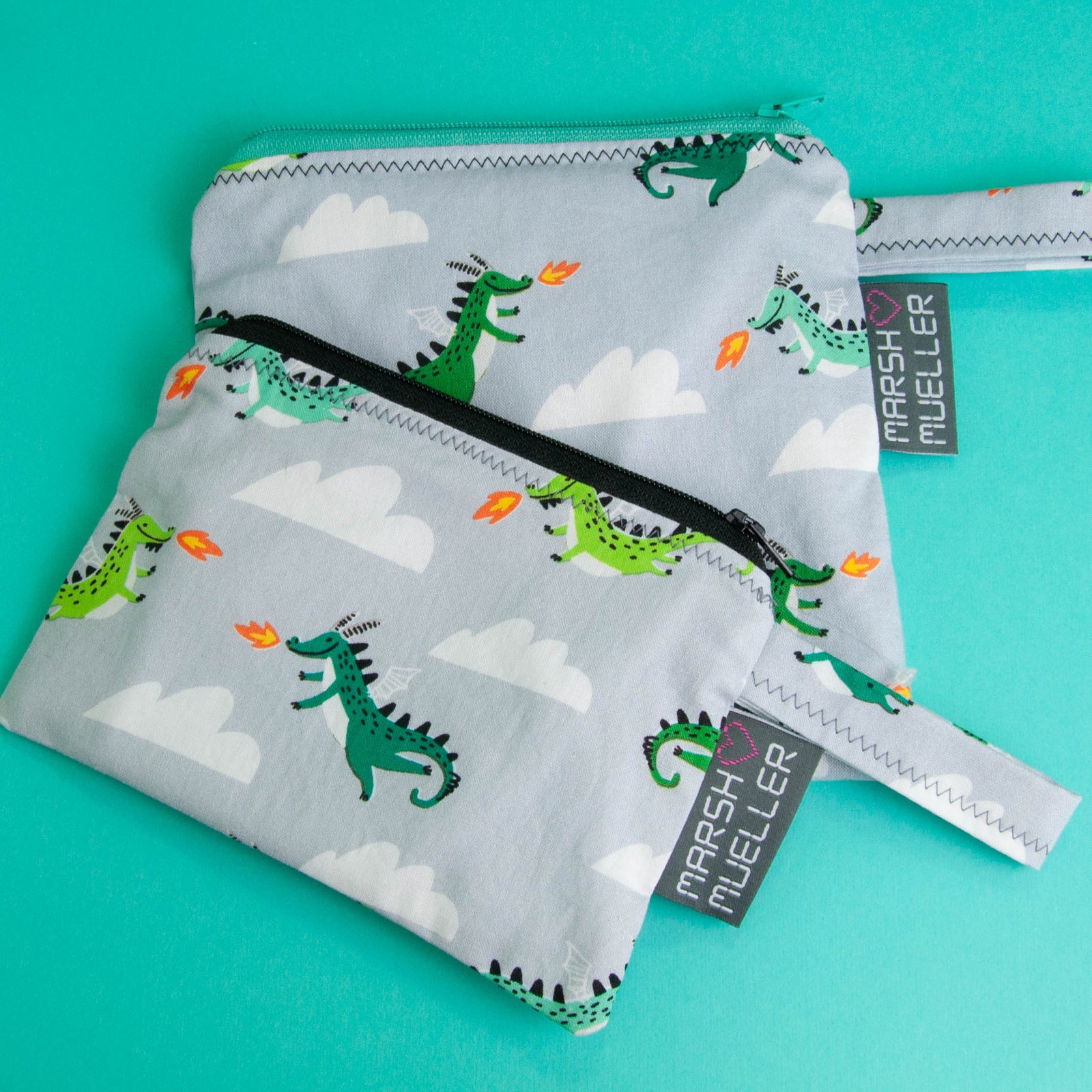 Image of Marshmueller Reusable Snack and Sandwich Bags arranged slightly overlapping in a flatlay image on a teal background. The bags are made from a dragons in the sky fabric print on a grey background.