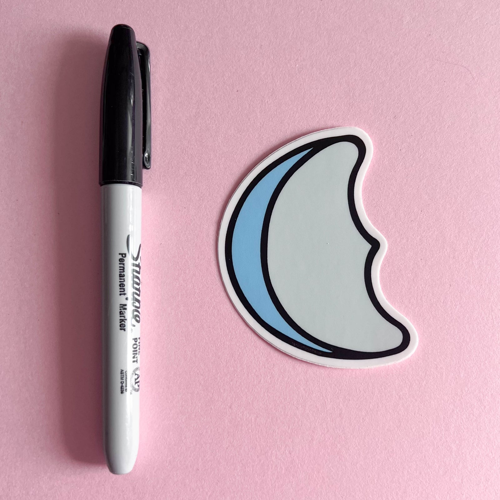 Moon-shaped sticker of blue marshmallow design next to a Sharpie marker on a pink background