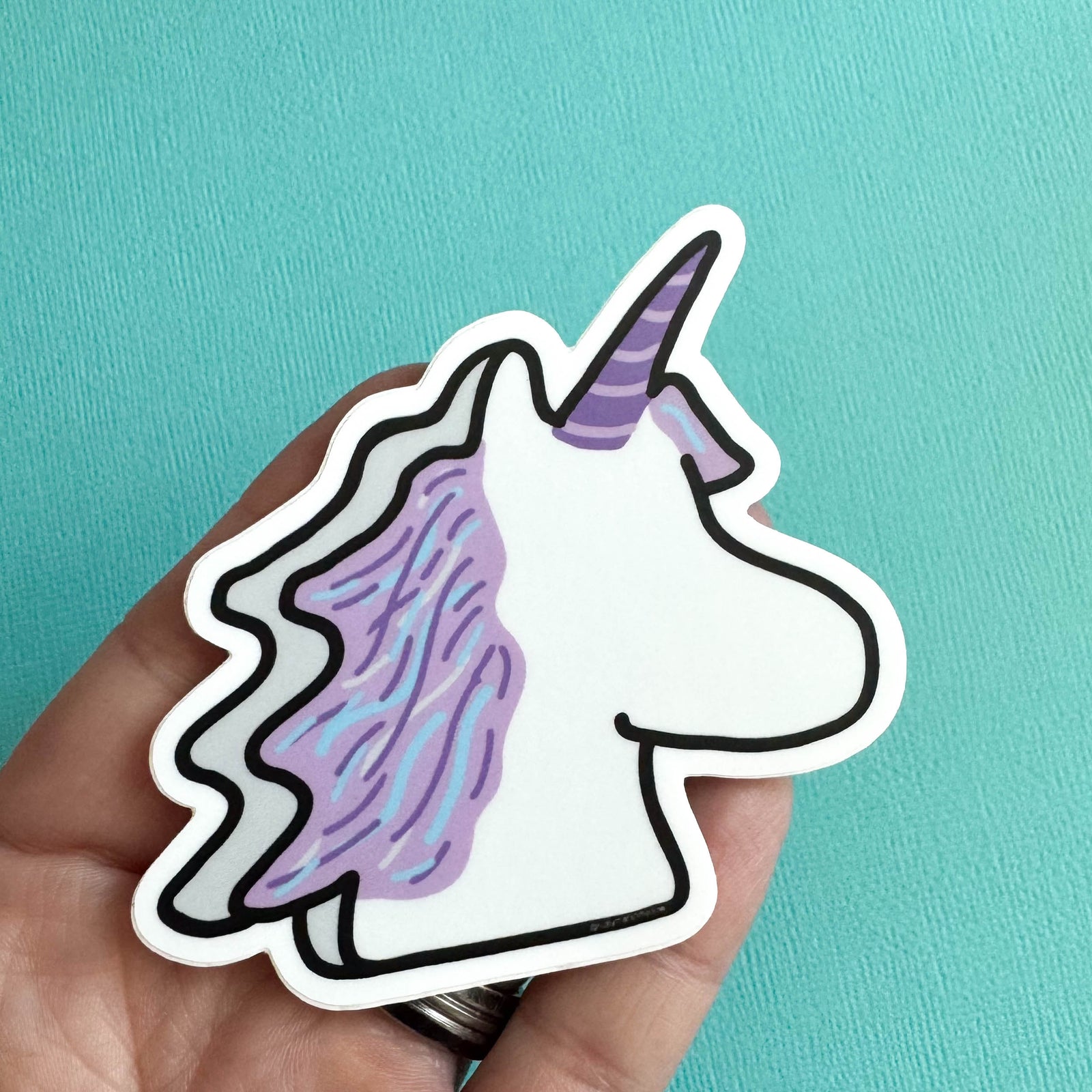 Hand holding a marshmallow unicorn sticker with a purple mane against a teal background