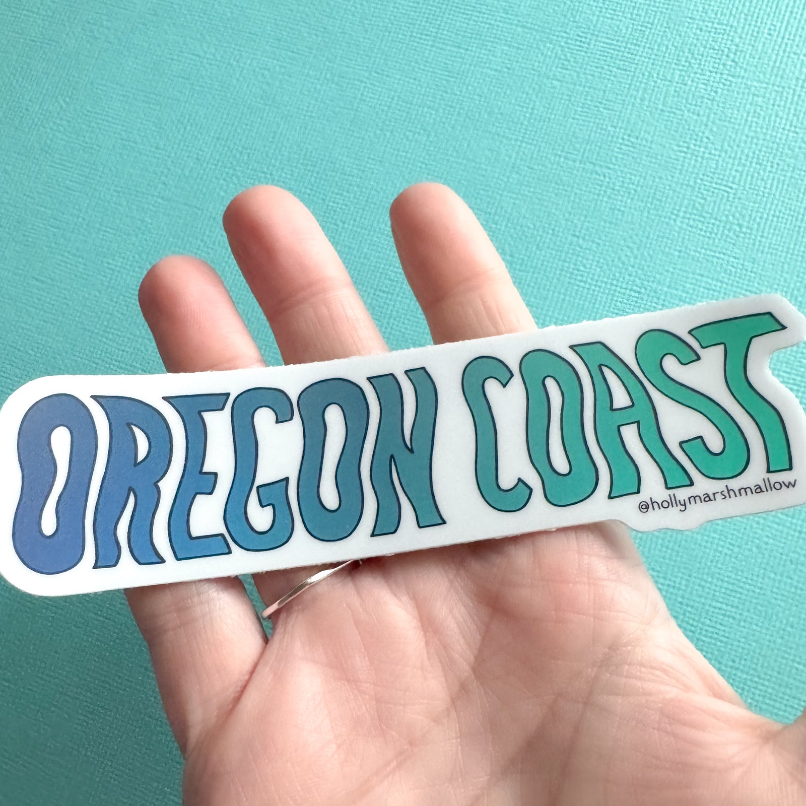 Hand holding an 'OREGON COAST' sticker with a teal background