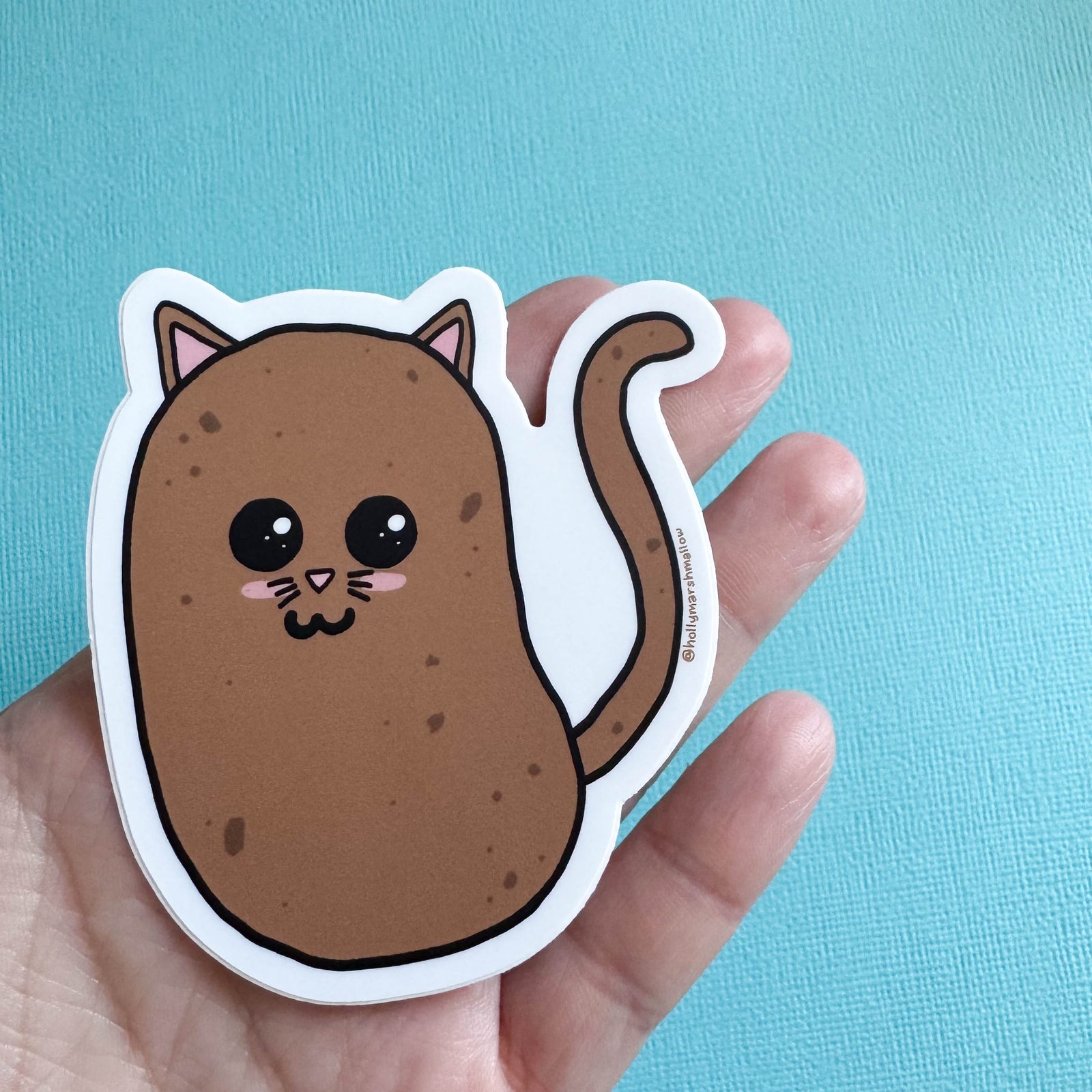Potato cat sticker held by a hand against a blue background