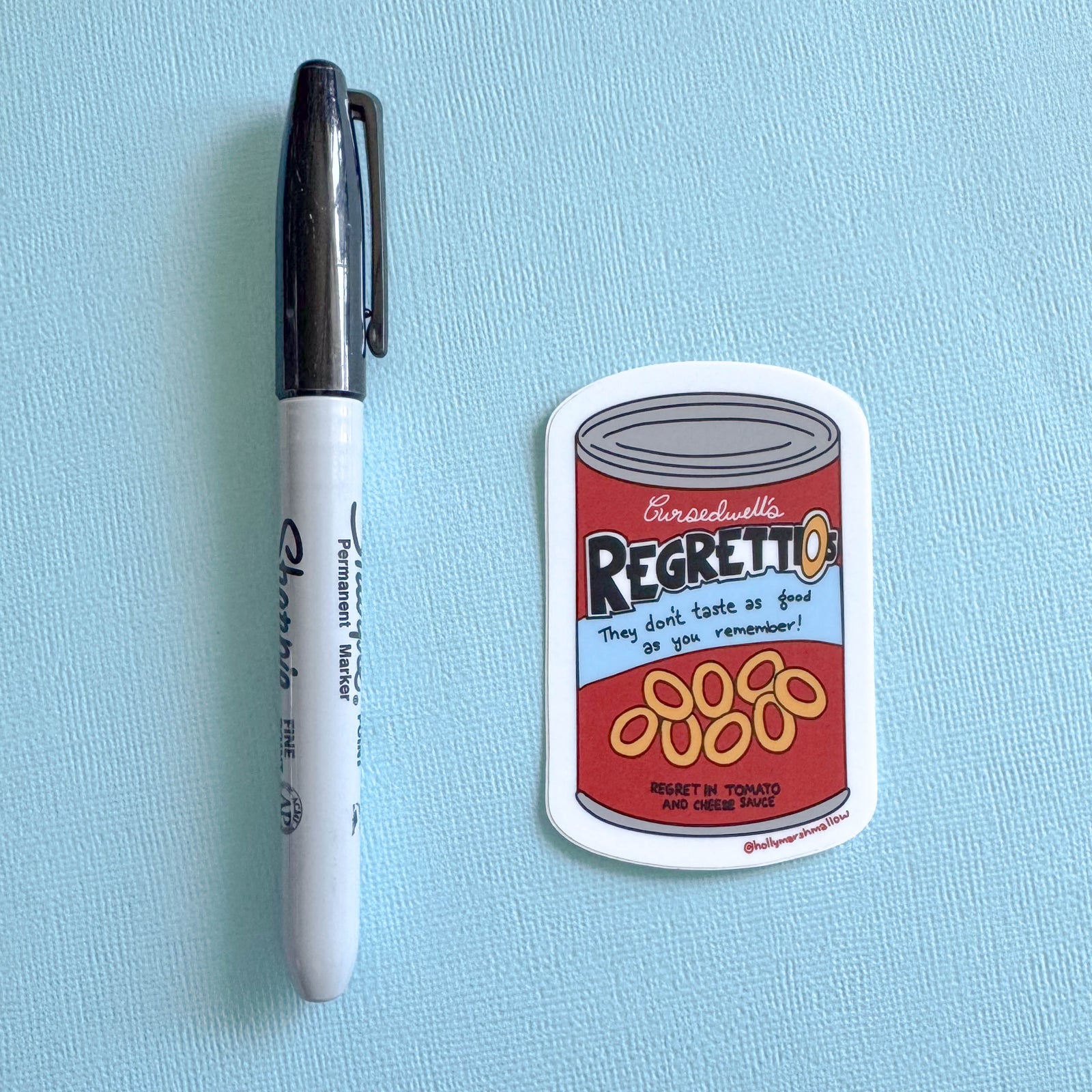 Sticker resembling a can labeled 'Regrettios' next to a pen on a light blue background