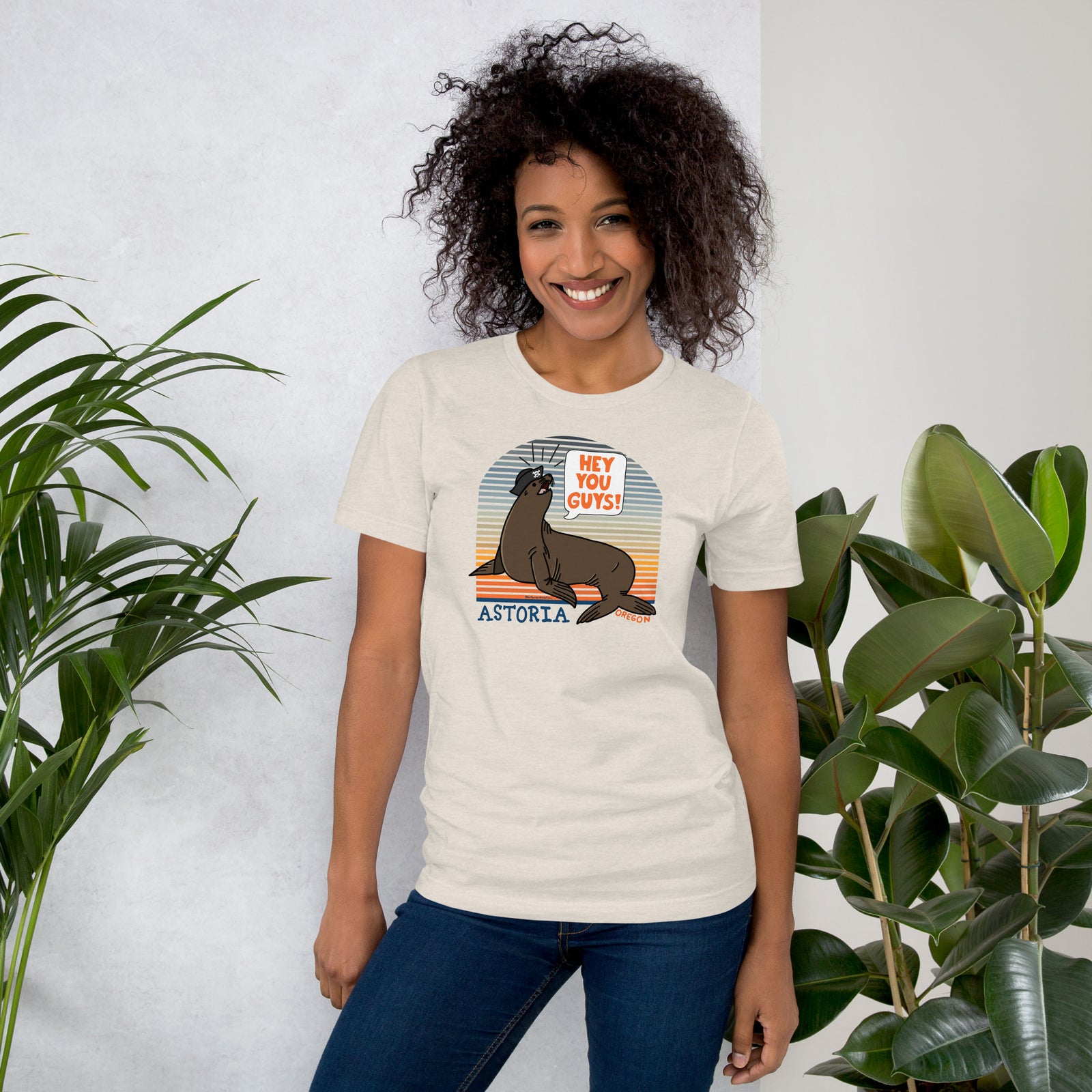 A black woman standing in front of a grey background with plants on both sides wearing jeans and an heather dust t-shirt of a sea lion in a tricorn hat yelling HEY YOU GUYS with a sunset stripe background and hand-lettered Astoria, Oregon on the bottom