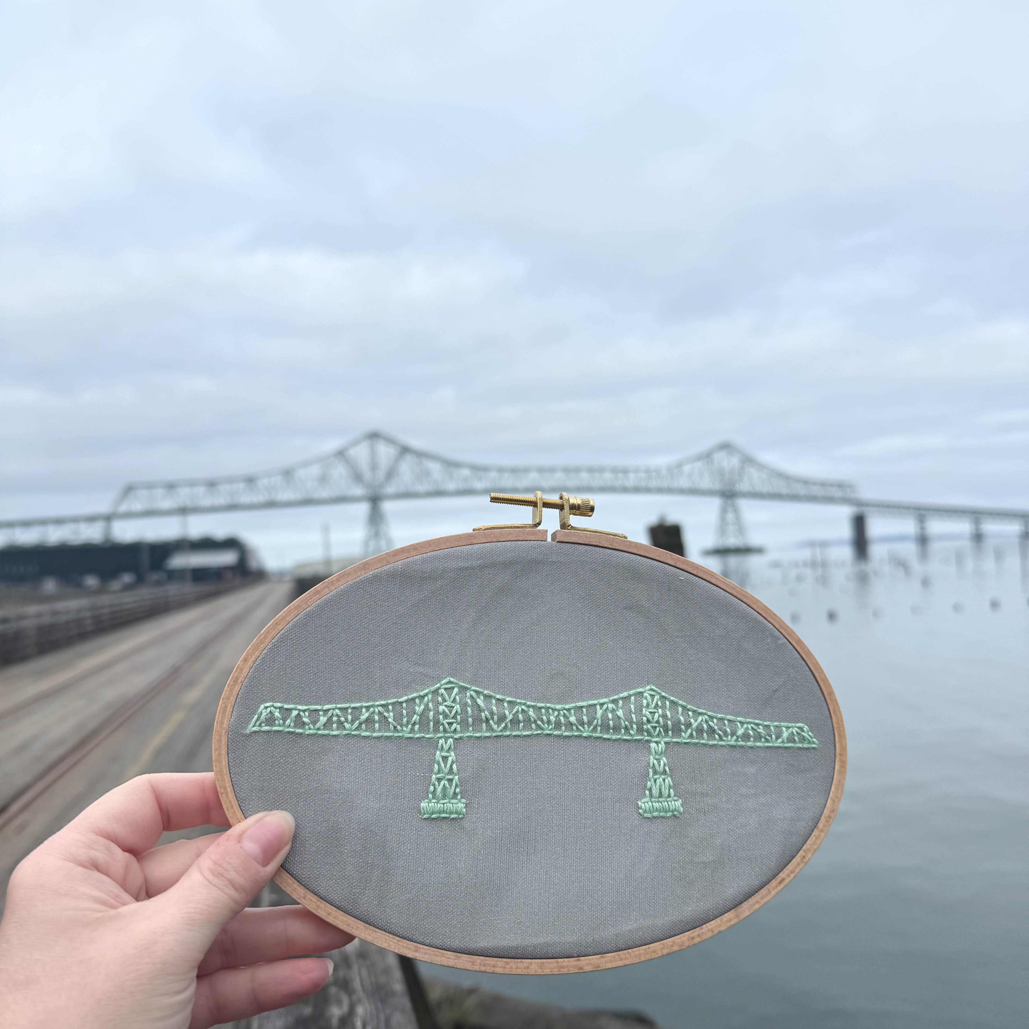 A hand holding a completed Megler Bridge Embroidery Kit on the Astoria Riverwalk with the Megler Bridge in the background