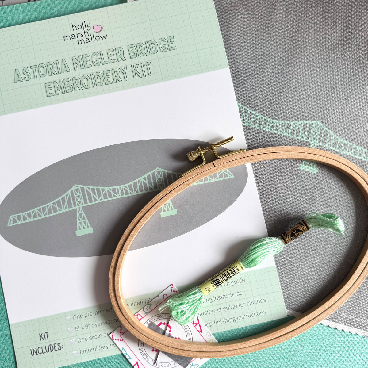 A flatlay of the contents of the Megler Bridge Embroidery Kit- the front insert, pre-printed fabric, oval embroidery hoop, a skein of embroidery floss, and a needle taped to a business card