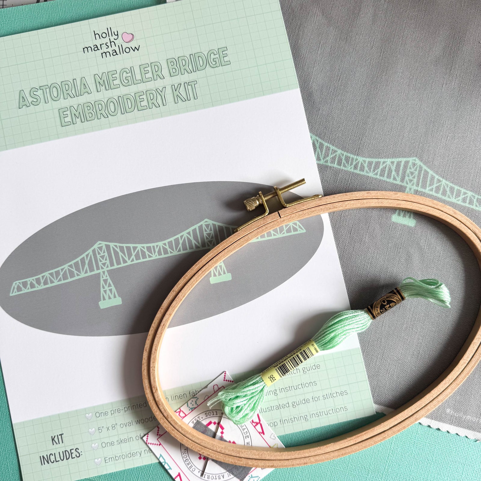 A flatlay of the contents of the Megler Bridge Embroidery Kit- the front insert, pre-printed fabric, oval embroidery hoop, a skein of embroidery floss, and a needle taped to a business card