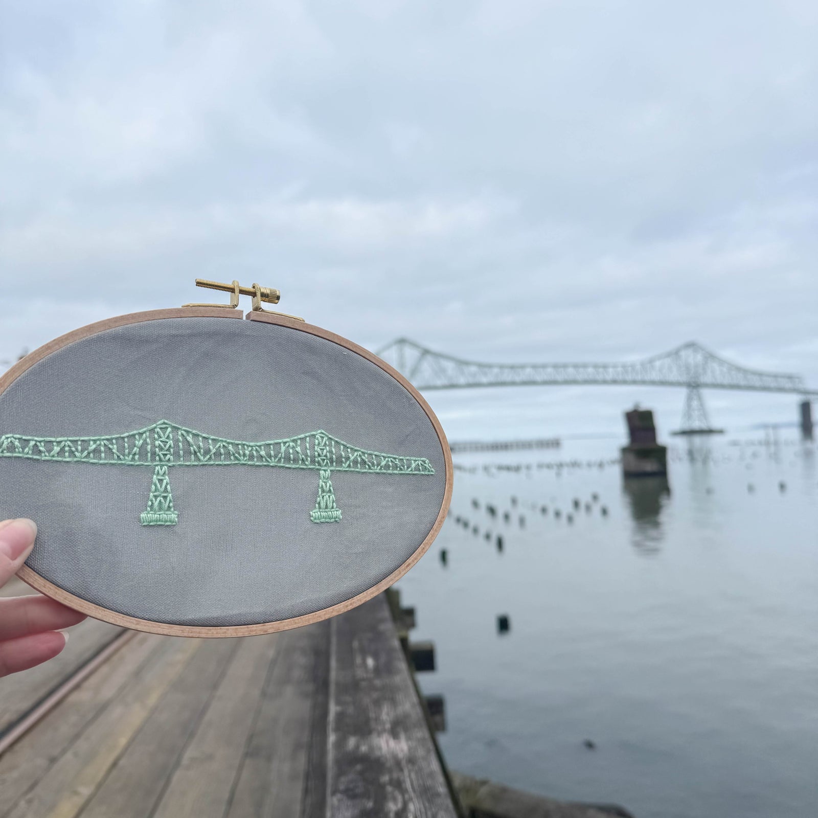 A hand holding a completed Megler Bridge Embroidery Kit on the Astoria Riverwalk with the Megler Bridge in the background