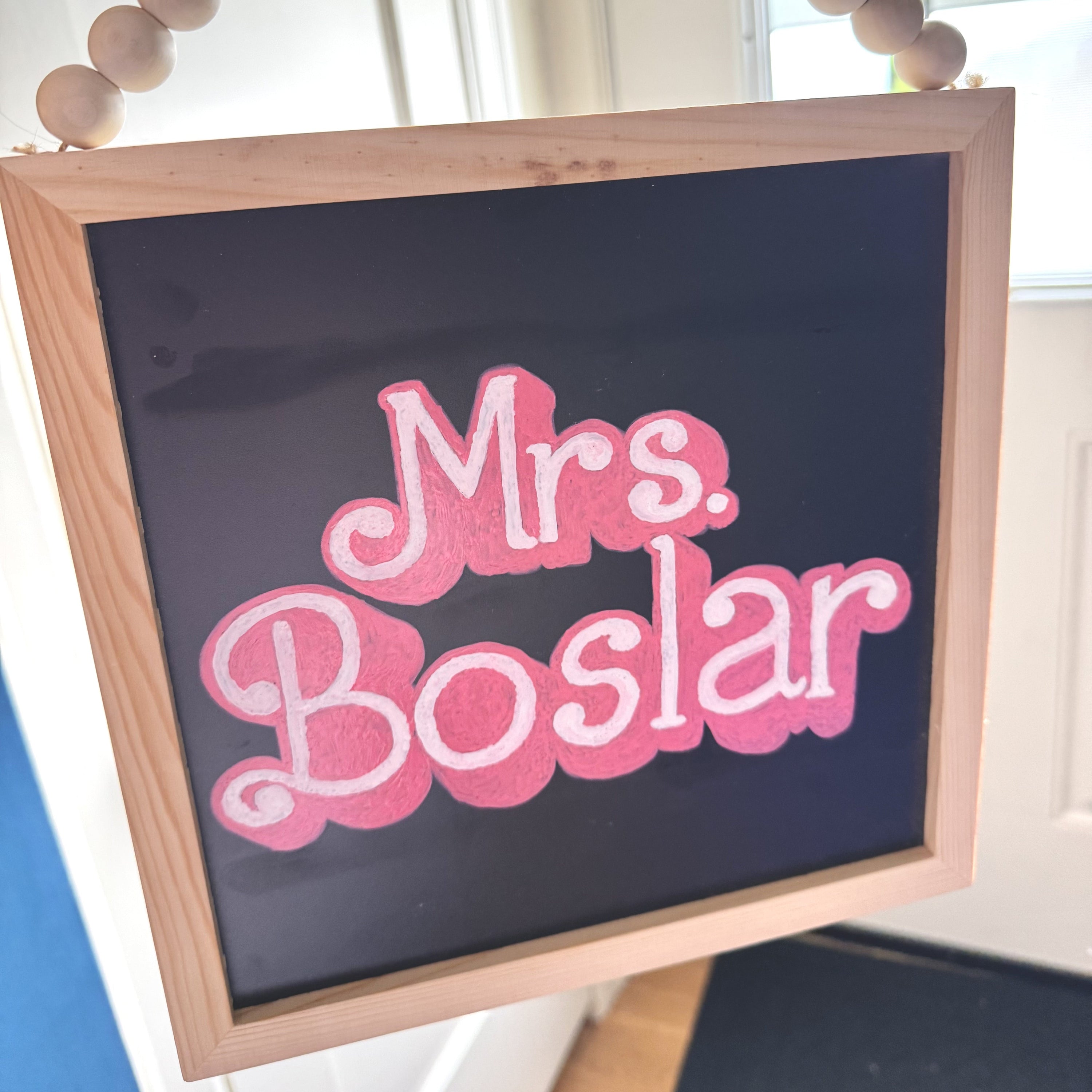 Chalkboard sign with 'Mrs. Boslar' in Barbie-inspired pink hand lettering, hanging on a wooden frame.