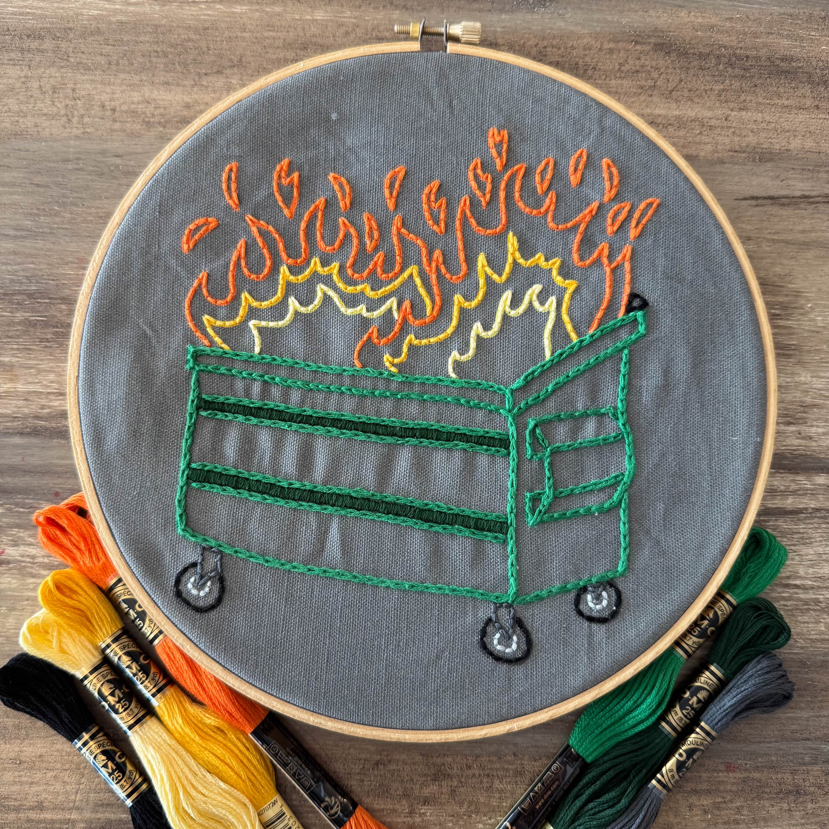 Embroidery hoop with a Dumpster Fire design on gray fabric, surrounded by embroidery threads.
