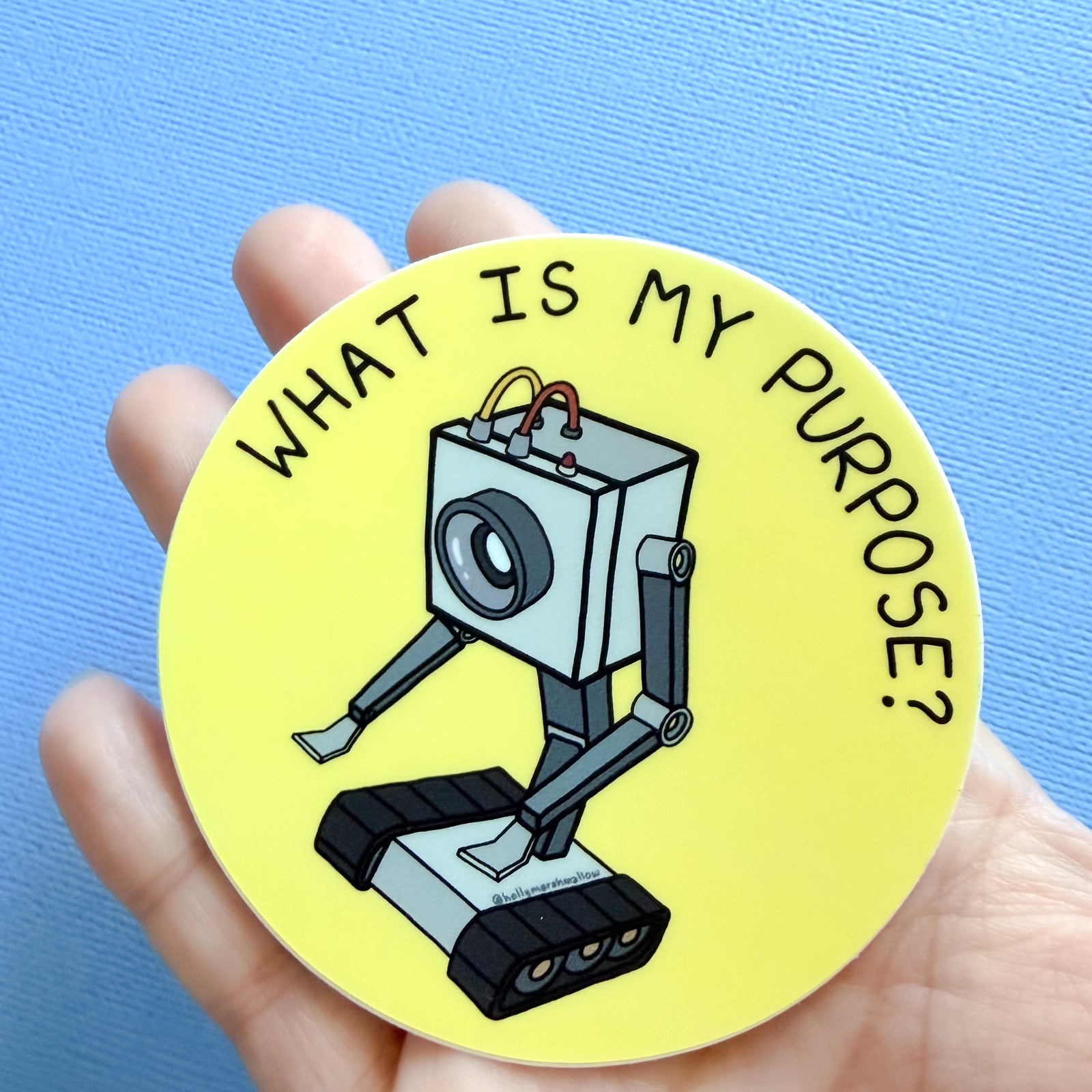 A hand holding a Yellow circle sticker with the Butter Robot from Rick + Morty in the center with hand-lettered text that reads What is My Purpose? on a blue background