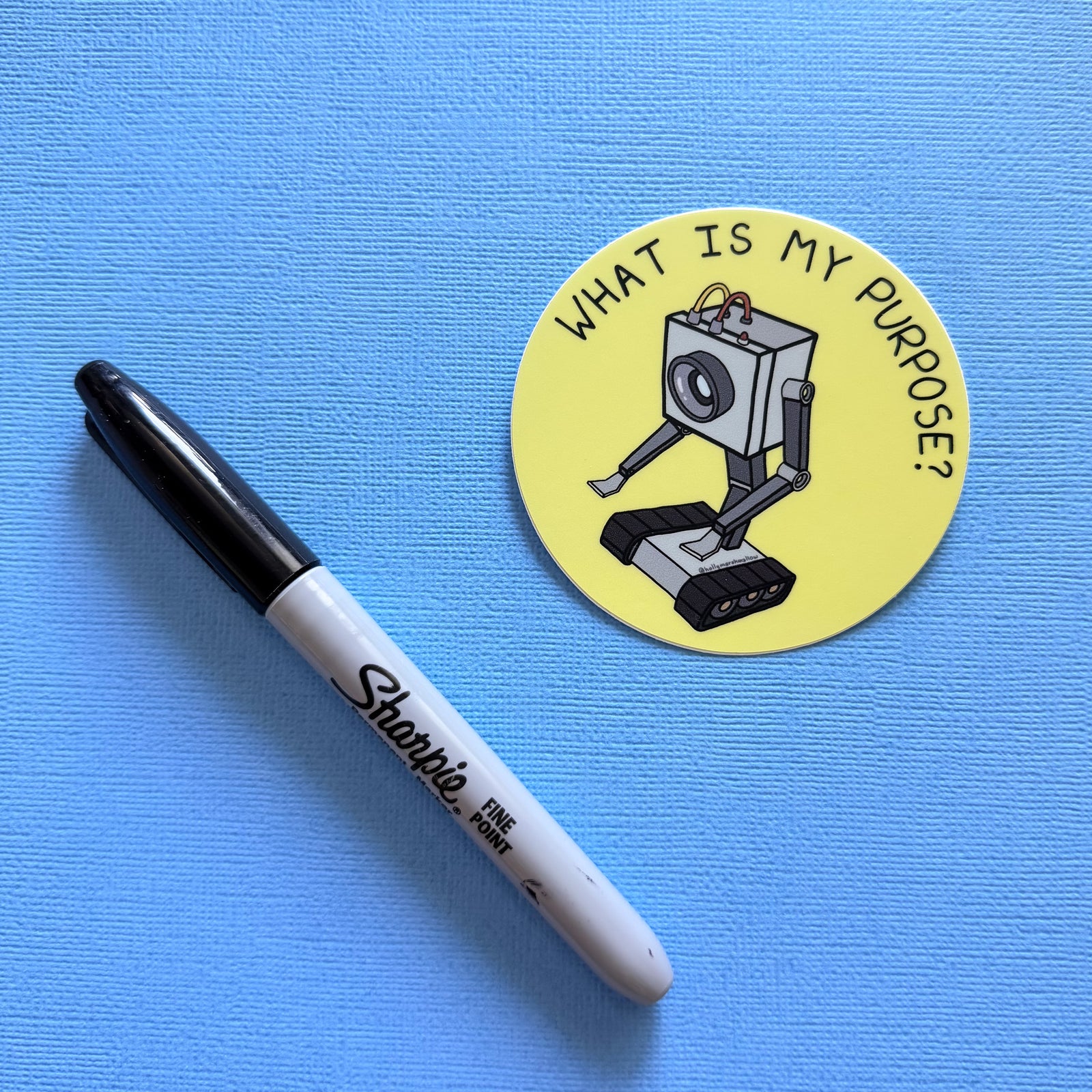 Yellow circle sticker with the Butter Robot from Rick + Morty in the center with hand-lettered text that reads What is My Purpose? next to a Sharpie on a blue background