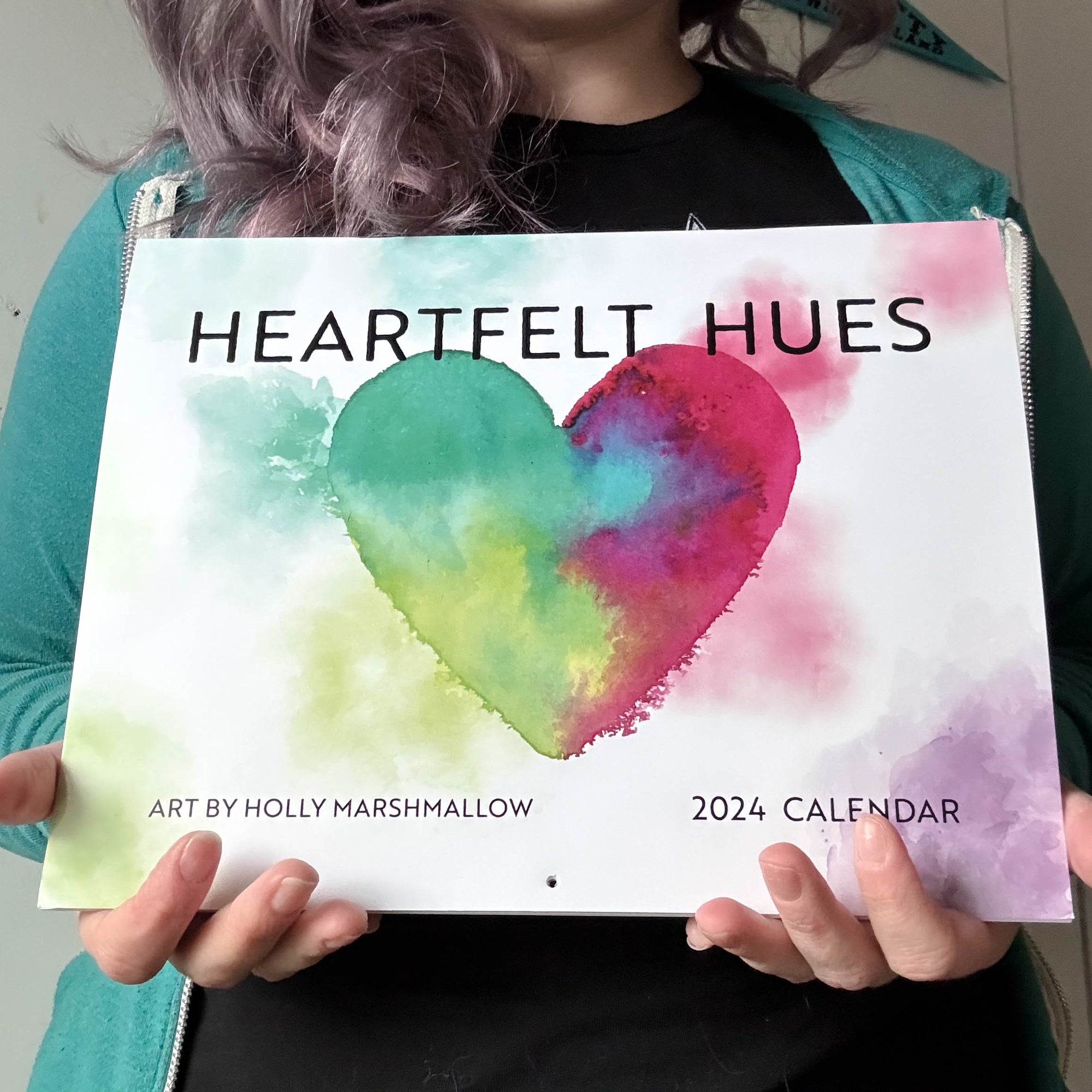 a white woman with purple hair and a teal hoodie holding the heartfelt hues calendar