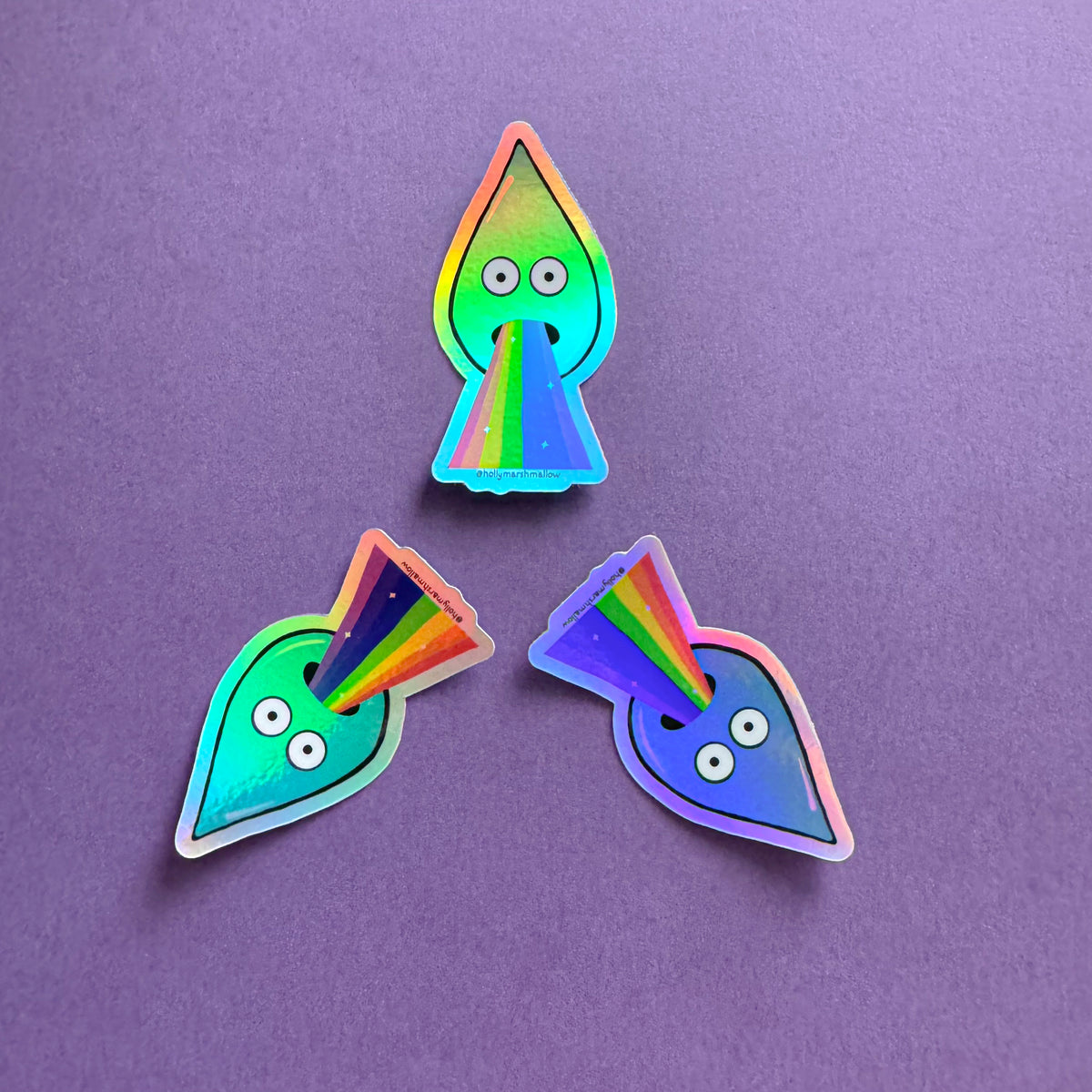 Trio of a sticker of a droplet with rainbow barf against a purple background