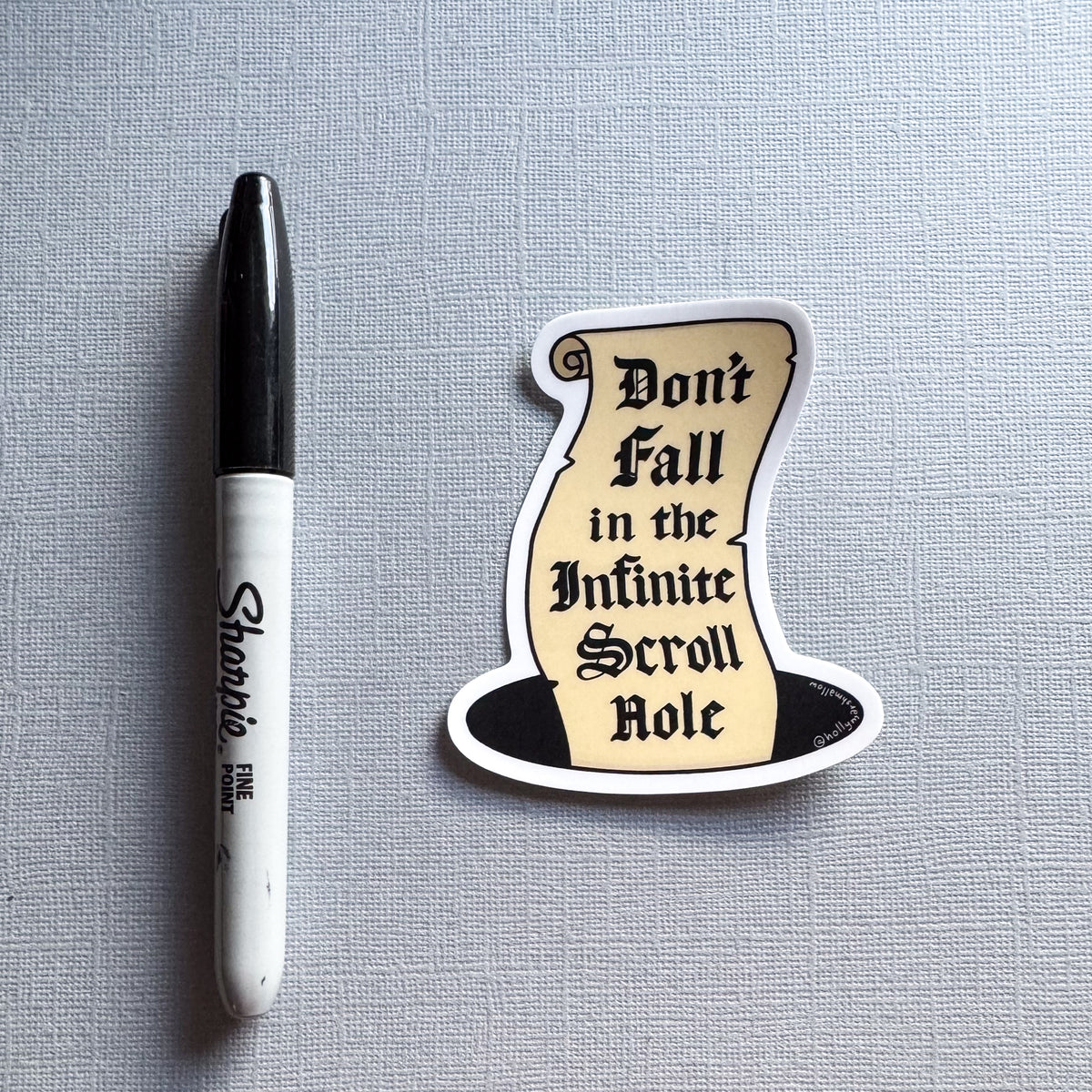 A die-cut sticker of an illustration of a beige scroll inside a hole, reading 'Don't Fall in the Infinite Scroll Role,' placed on a light gray textured surface next to a black and white Sharpie marker for scale.