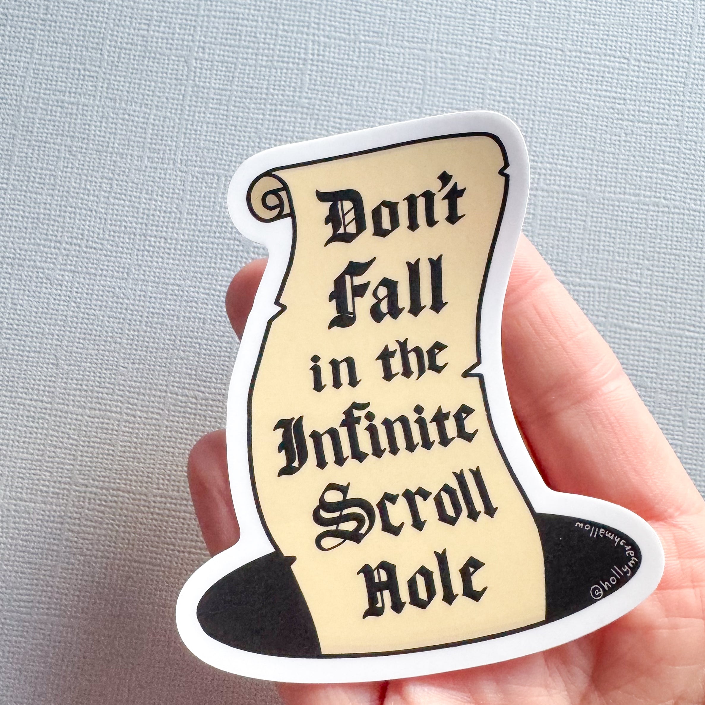A hand holding a die-cut sticker shaped like a black top hat with a parchment scroll insert that reads 'Don't Fall in the Infinite Scroll Role' in black gothic lettering. The sticker has a white border and is photographed against a light gray textured background.