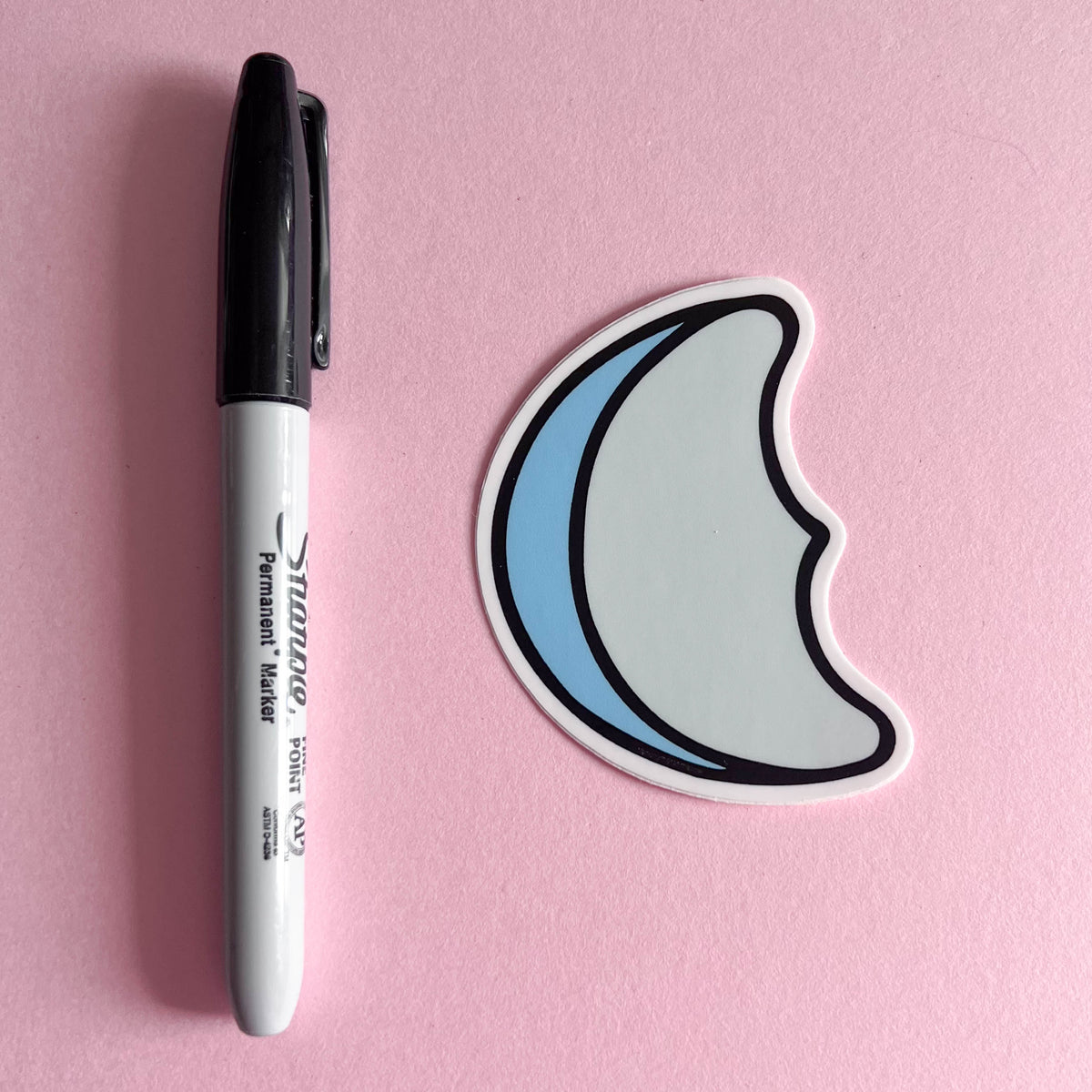 Moon-shaped sticker of blue marshmallow design next to a Sharpie marker on a pink background