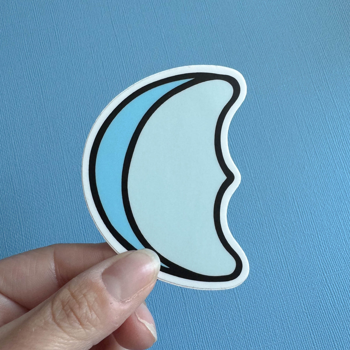 Hand holding a blue marshmallow crescent moon sticker against a blue background