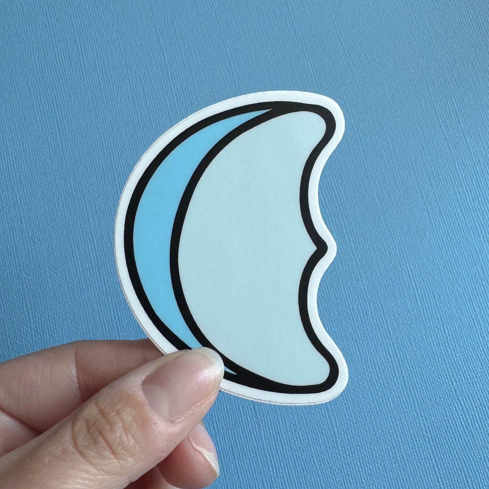 Hand holding a blue marshmallow crescent moon sticker against a blue background