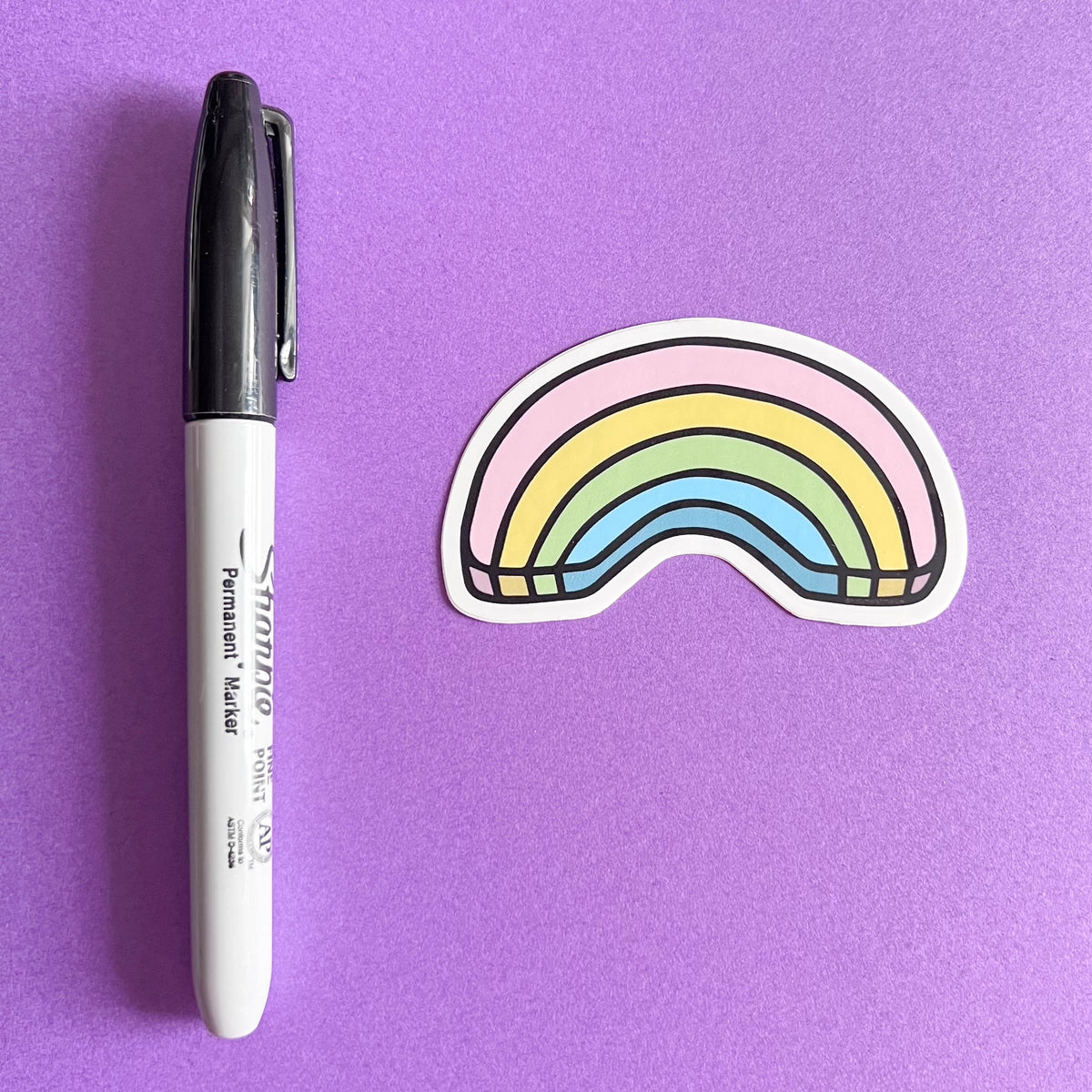 Rainbow-shaped sticker next to a Sharpie marker on a purple background