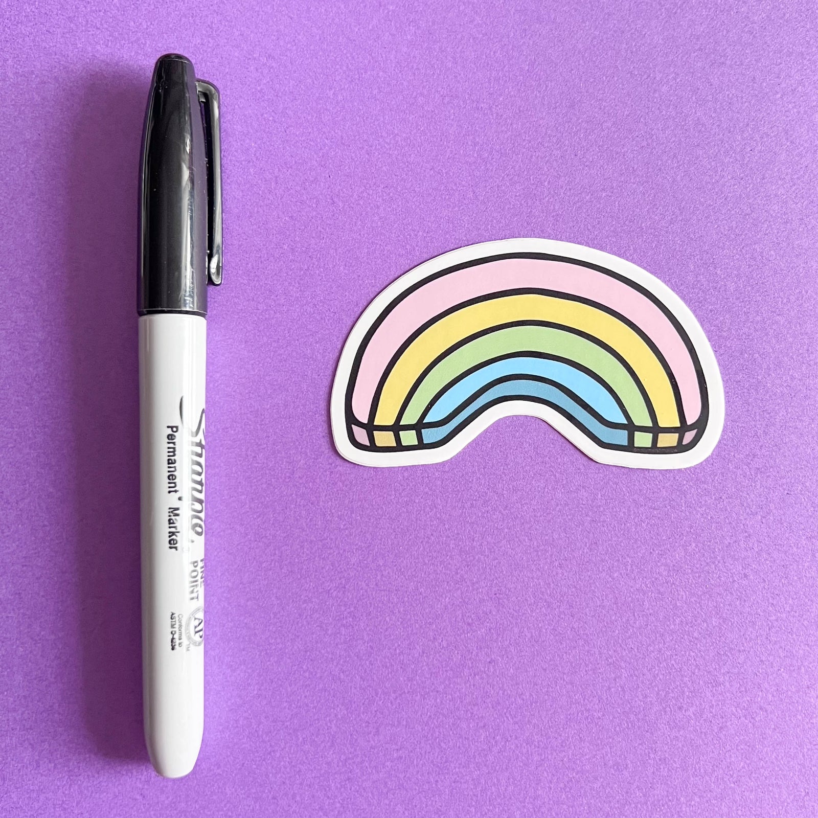 Rainbow-shaped sticker next to a Sharpie marker on a purple background