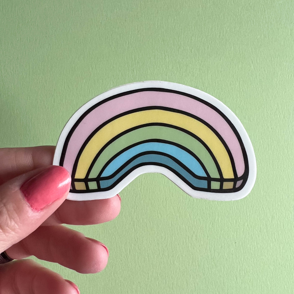 Hand holding a rainbow-shaped sticker against a light green background
