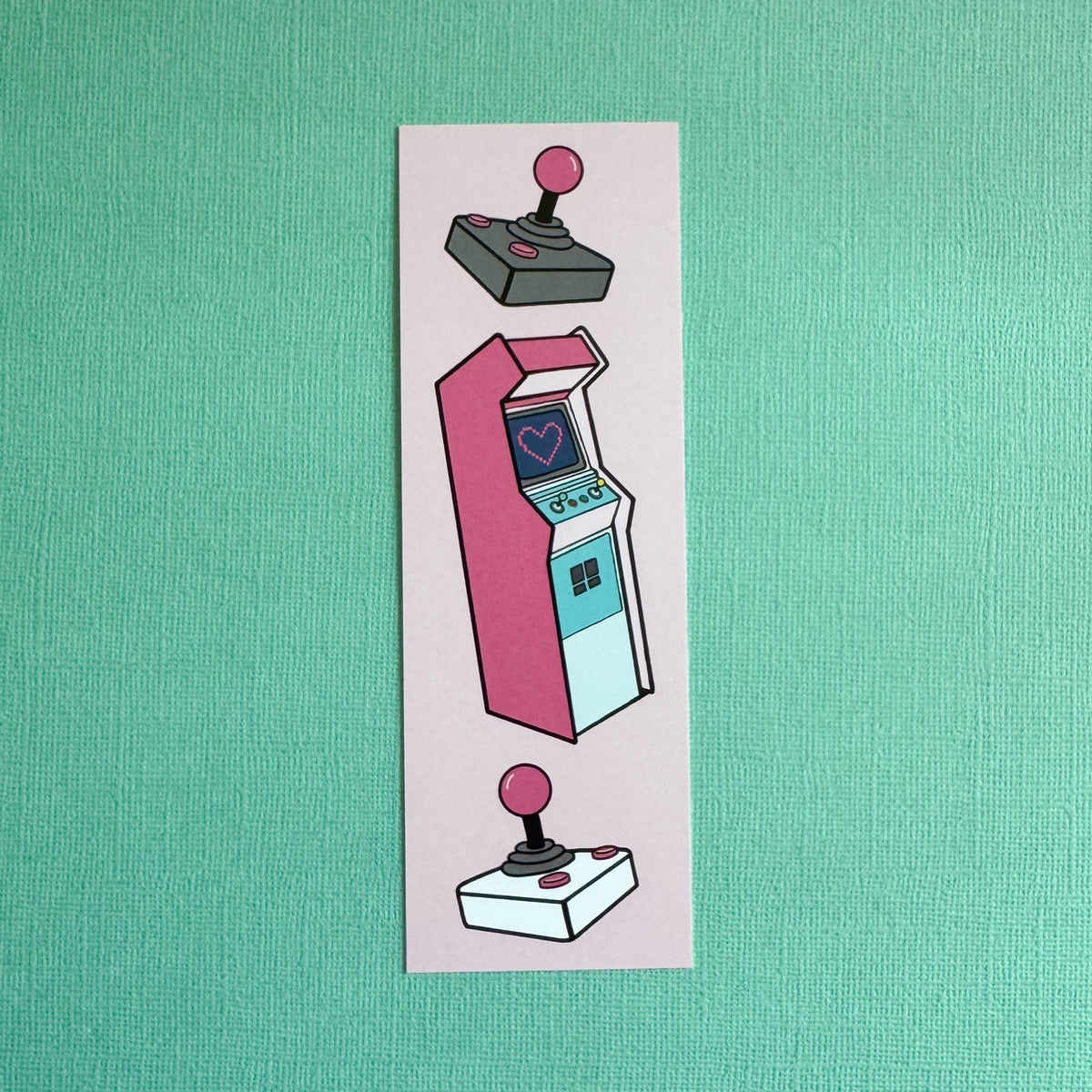 Front of bookmark with an illustration of a pink arcade machine with two joysticks on a pink background. Bookmark is laying on a solid teal background.