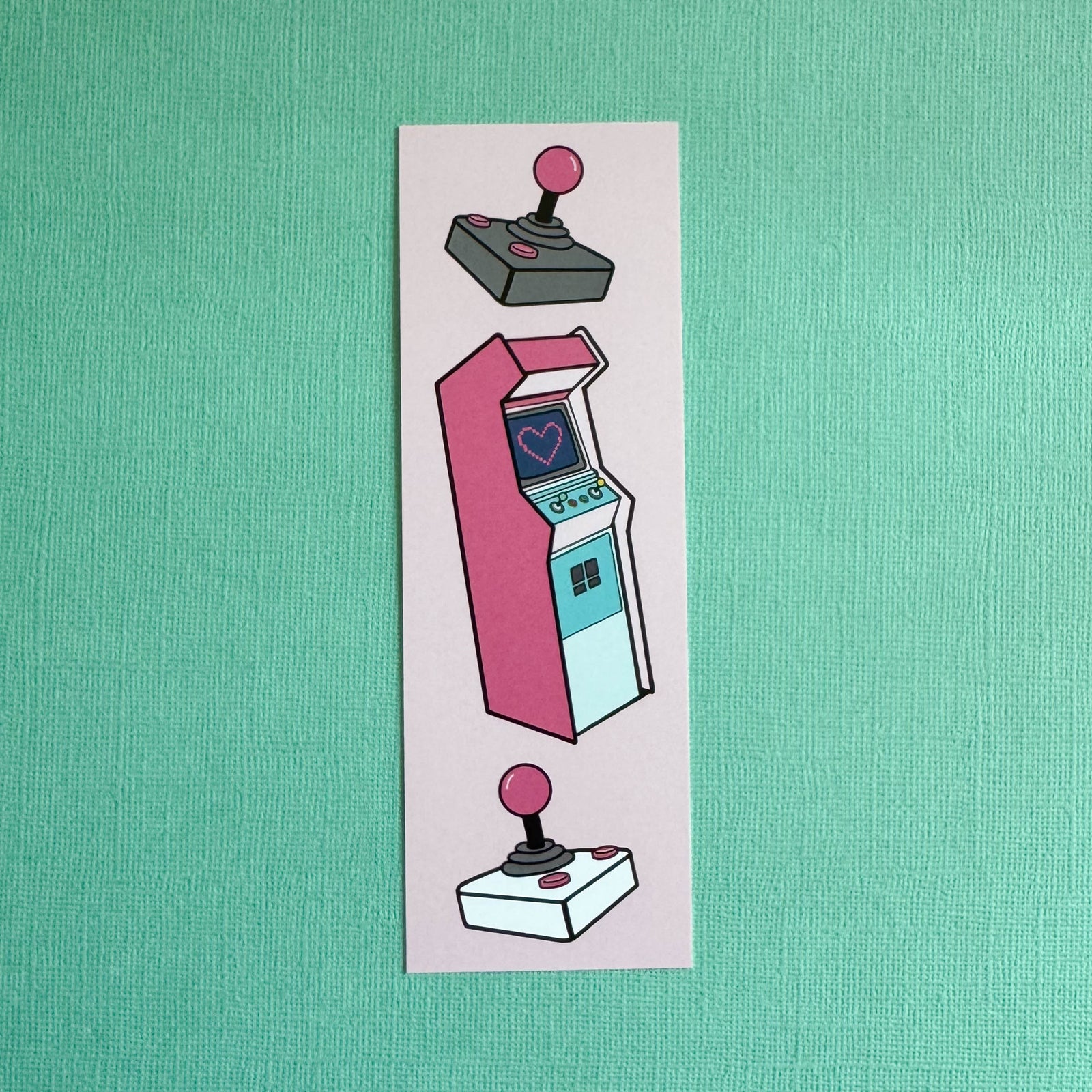 Front of bookmark with an illustration of a pink arcade machine with two joysticks on a pink background. Bookmark is laying on a solid teal background.