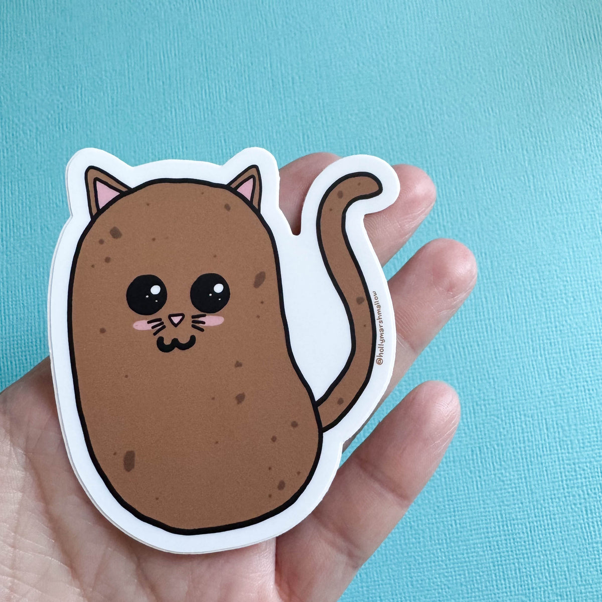 Potato cat sticker held by a hand against a blue background