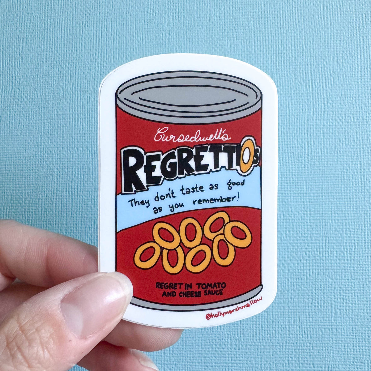 Sticker of a can labeled 'Cursedwell's Regrettios' held against a blue background