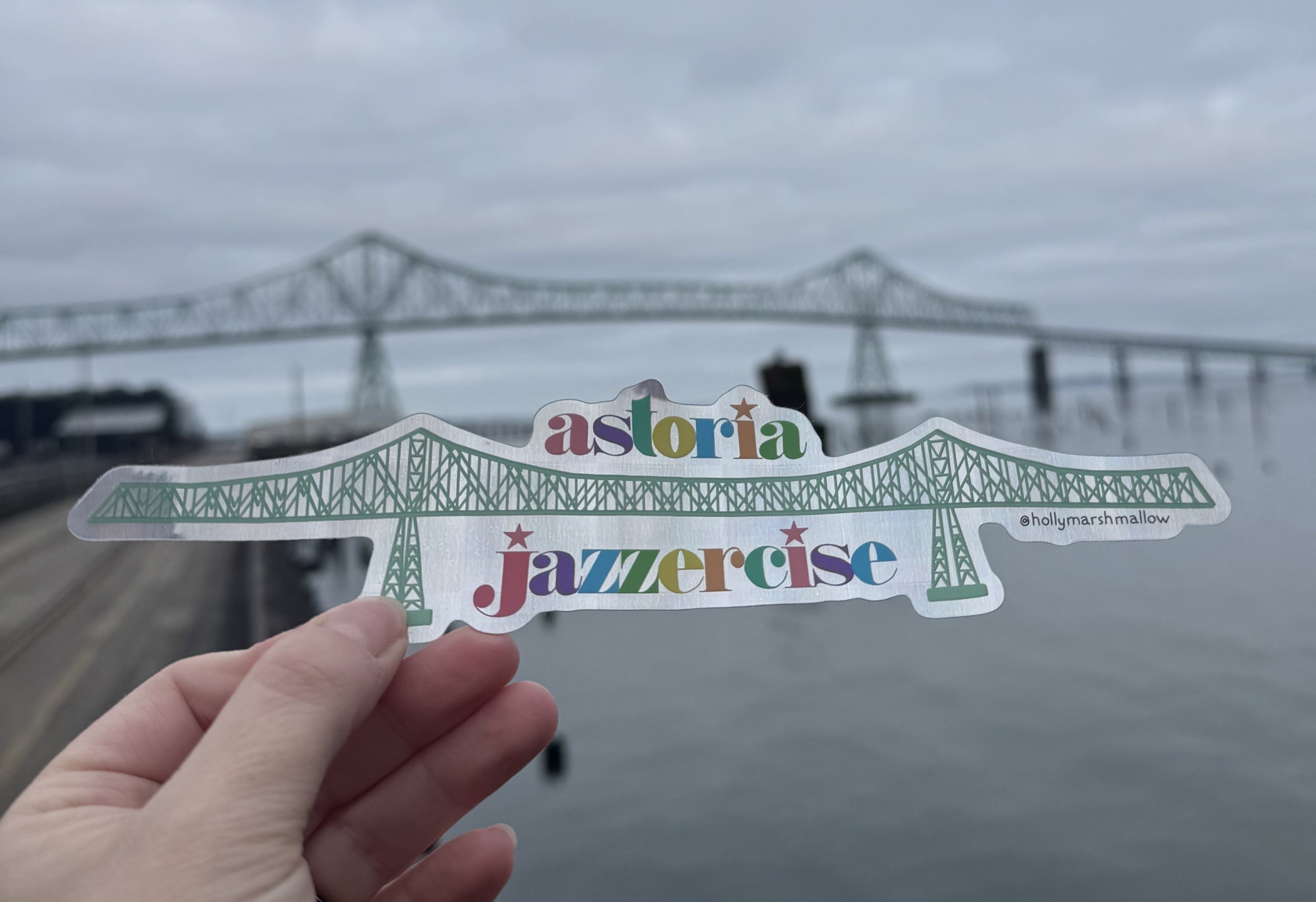 Hand holding a 'Astoria Jazzercise' sticker with a bridge in the background