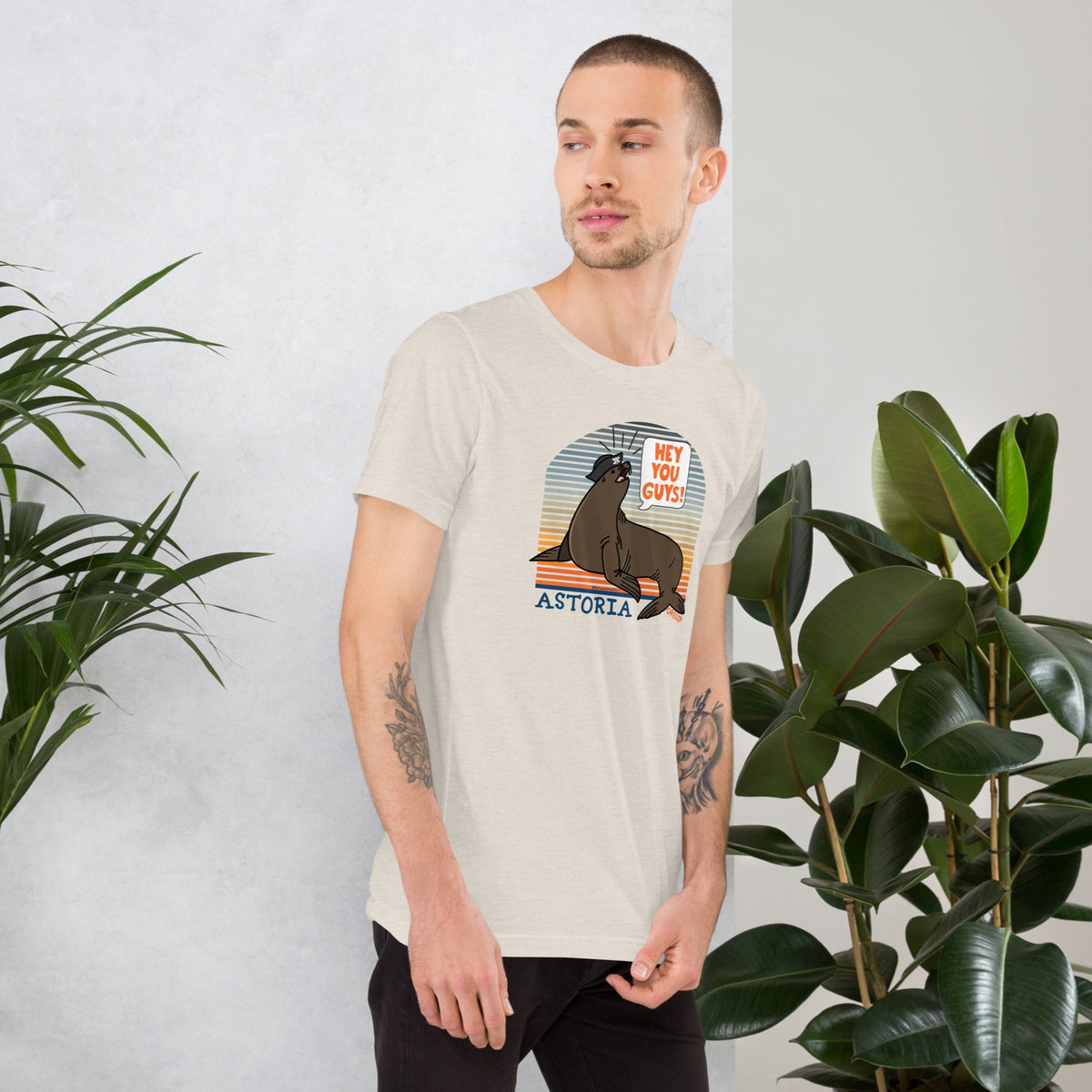 A white man stands in front of a light grey background wearing black pants and an ice blue t-shirt of a sea lion in a tricorn hat yelling HEY YOU GUYS with a sunset stripe background and hand-lettered Astoria, Oregon on the bottom