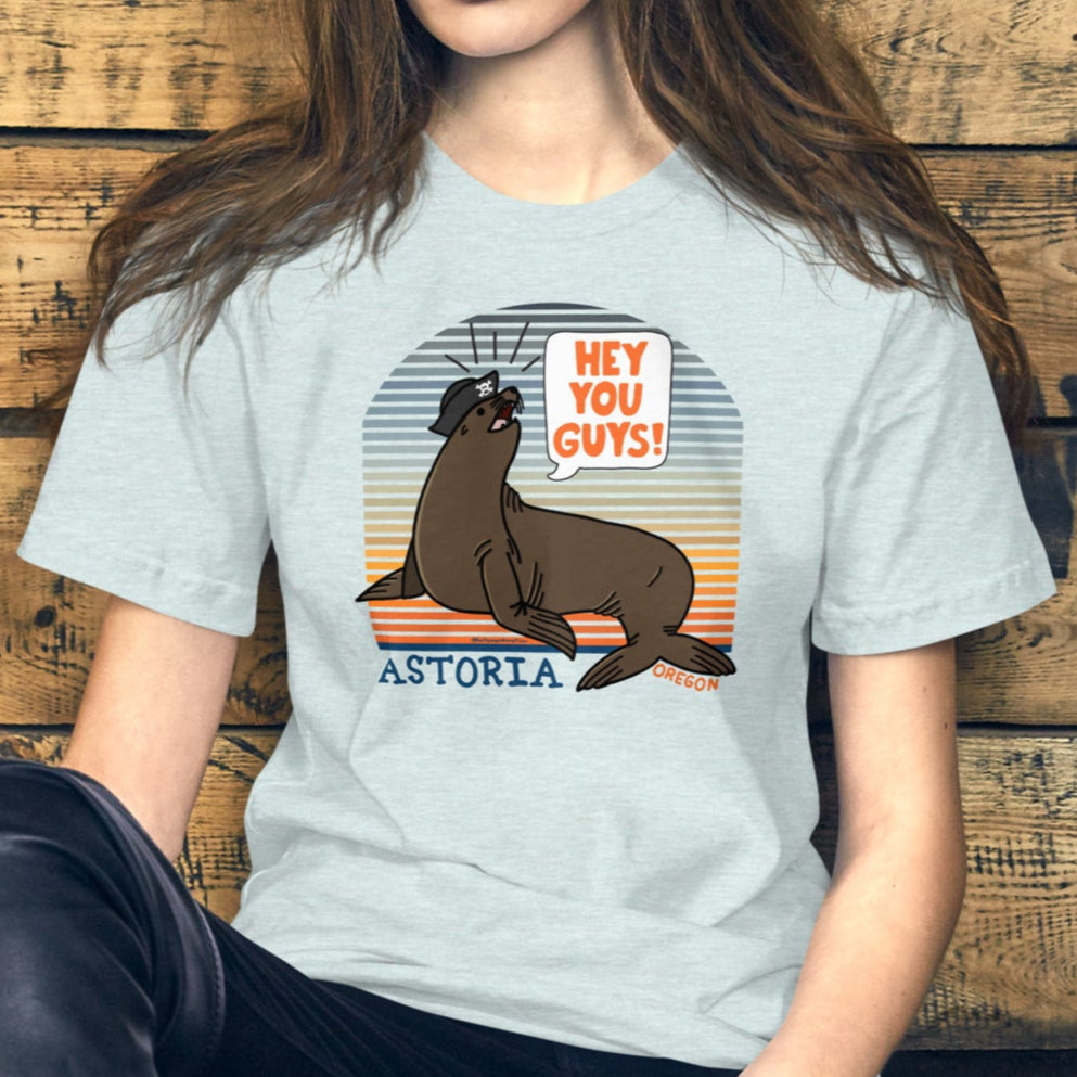 A white woman with long brunette hair leaning against a wooden slatwall background wearing an ice blue t-shirt of a sea lion in a tricorn hat yelling HEY YOU GUYS with a sunset stripe background and hand-lettered Astoria, Oregon on the bottom