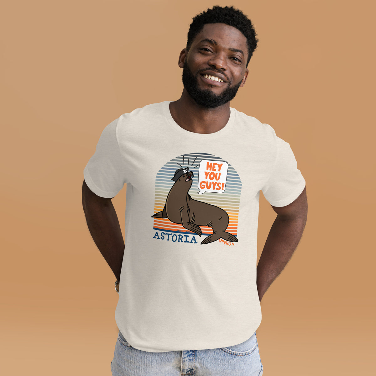 A black man syanding in front of a tan background wearing an heather dust t-shirt of a sea lion in a tricorn hat yelling HEY YOU GUYS with a sunset stripe background and hand-lettered Astoria, Oregon on the bottom