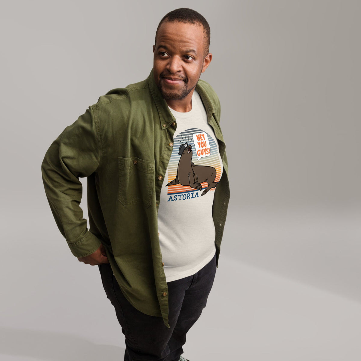 A black man standing in front of a grey sweep background wearing black pants, an olive green button up shirt, open to reveal an ice blue t-shirt of a sea lion in a tricorn hat yelling HEY YOU GUYS with a sunset stripe background and hand-lettered Astoria, Oregon on the bottom