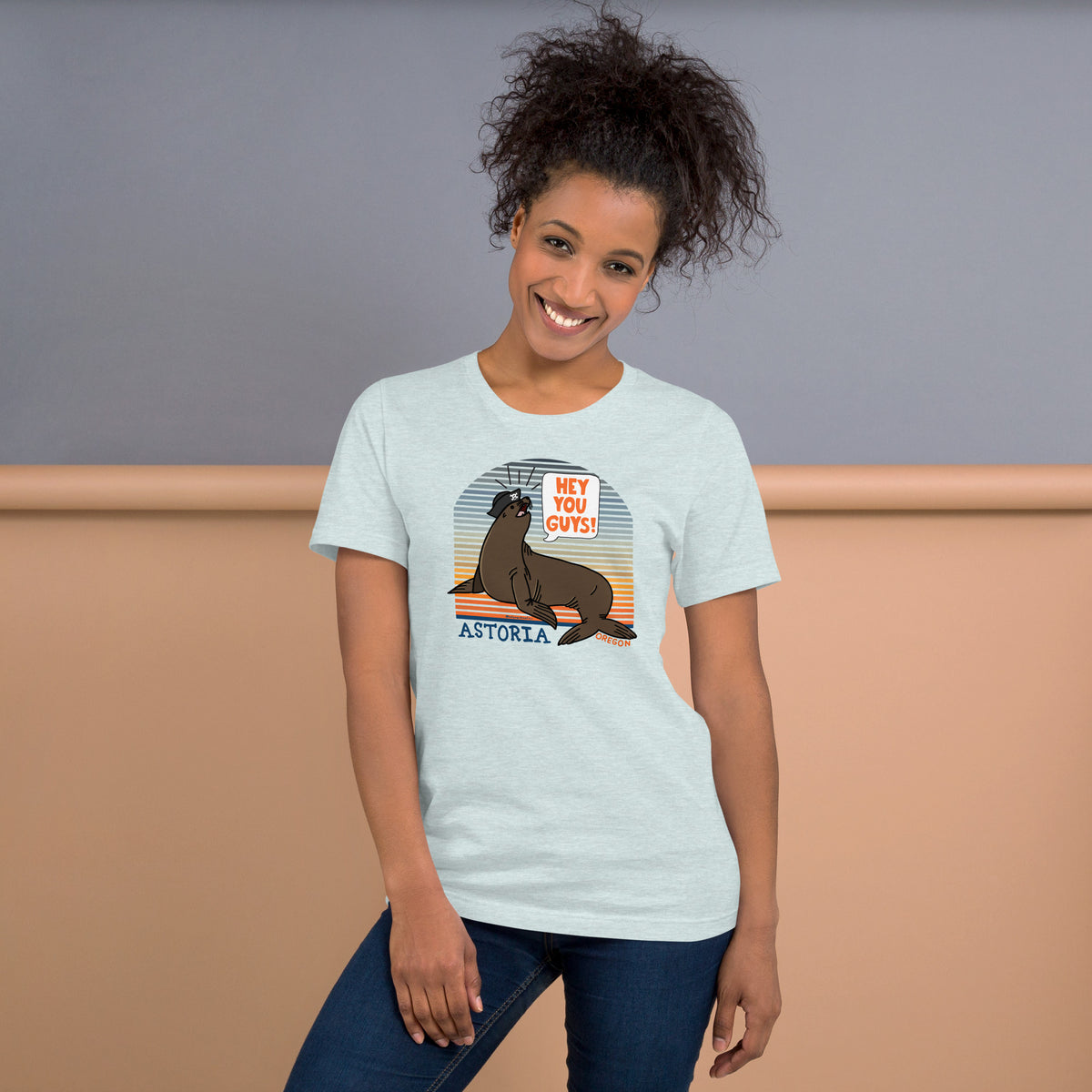A black woman stands in front of a grey and wood background wearing jeans and an ice blue t-shirt of a sea lion in a tricorn hat yelling HEY YOU GUYS with a sunset stripe background and hand-lettered Astoria, Oregon on the bottom