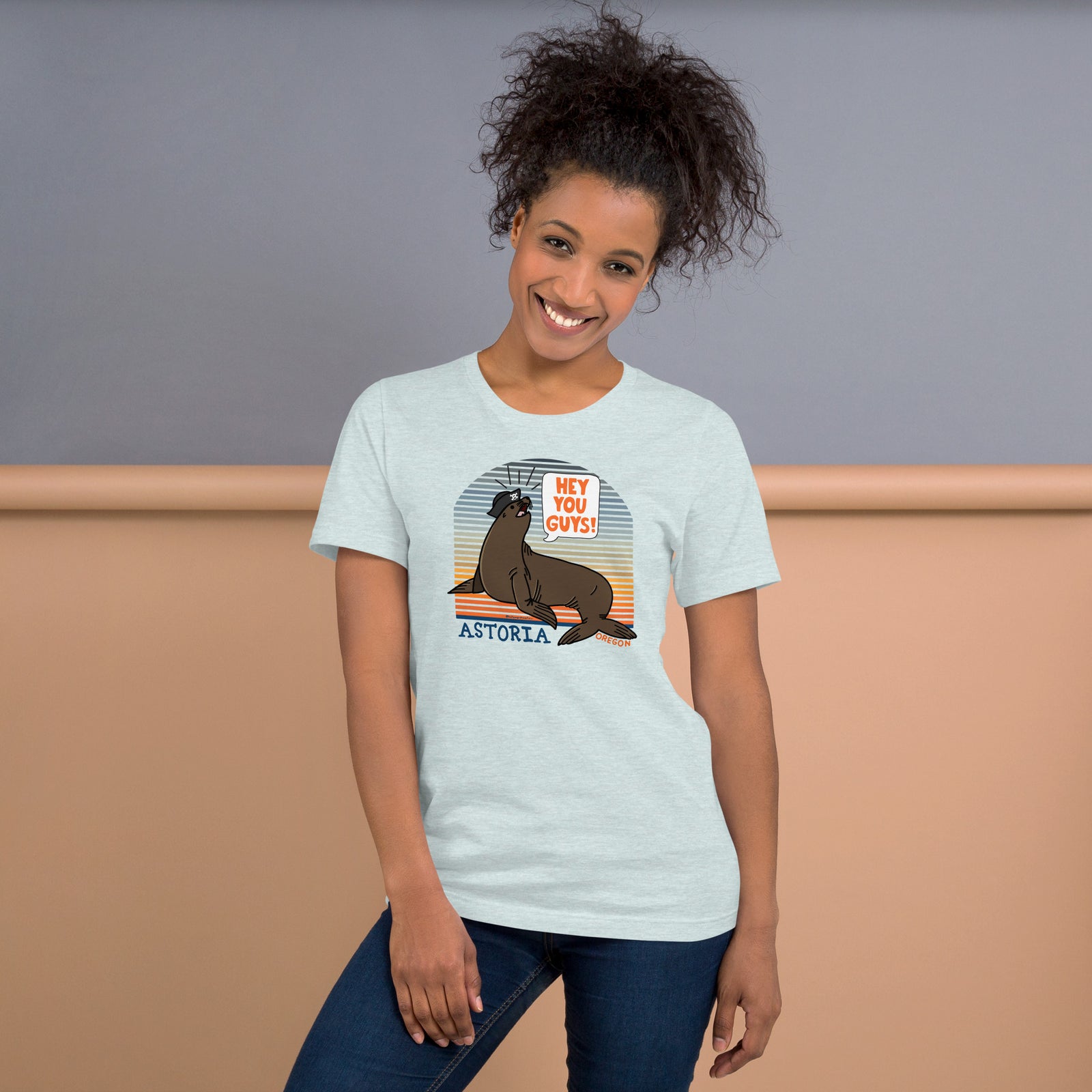 A black woman stands in front of a grey and wood background wearing jeans and an ice blue t-shirt of a sea lion in a tricorn hat yelling HEY YOU GUYS with a sunset stripe background and hand-lettered Astoria, Oregon on the bottom
