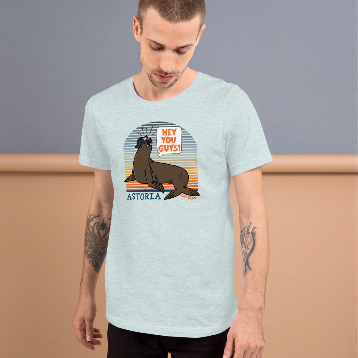 A white man standing in front of a grey and wood background wearing an ice blue t-shirt of a sea lion in a tricorn hat yelling HEY YOU GUYS with a sunset stripe background and hand-lettered Astoria, Oregon on the bottom