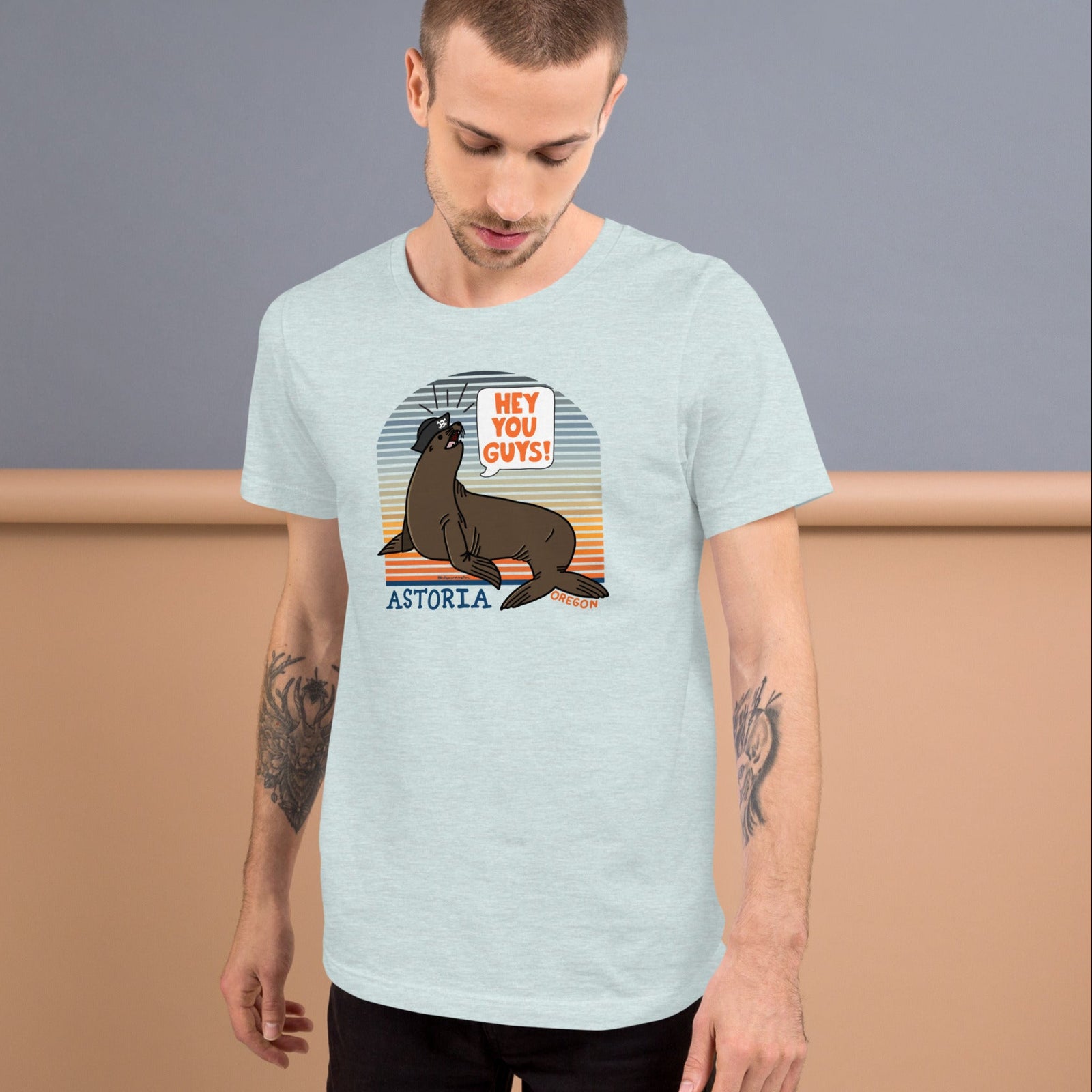 A white man standing in front of a grey and wood background wearing an ice blue t-shirt of a sea lion in a tricorn hat yelling HEY YOU GUYS with a sunset stripe background and hand-lettered Astoria, Oregon on the bottom