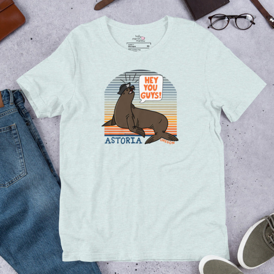 A flatlay of an ice blue t-shirt of a sea lion in a tricorn hat yelling HEY YOU GUYS with a sunset stripe background and hand-lettered Astoria, Oregon on the bottom. A pair of jeans lays to the bottom left, eyeglasses to the upper right, and a pair of shoes to the bottom right.
