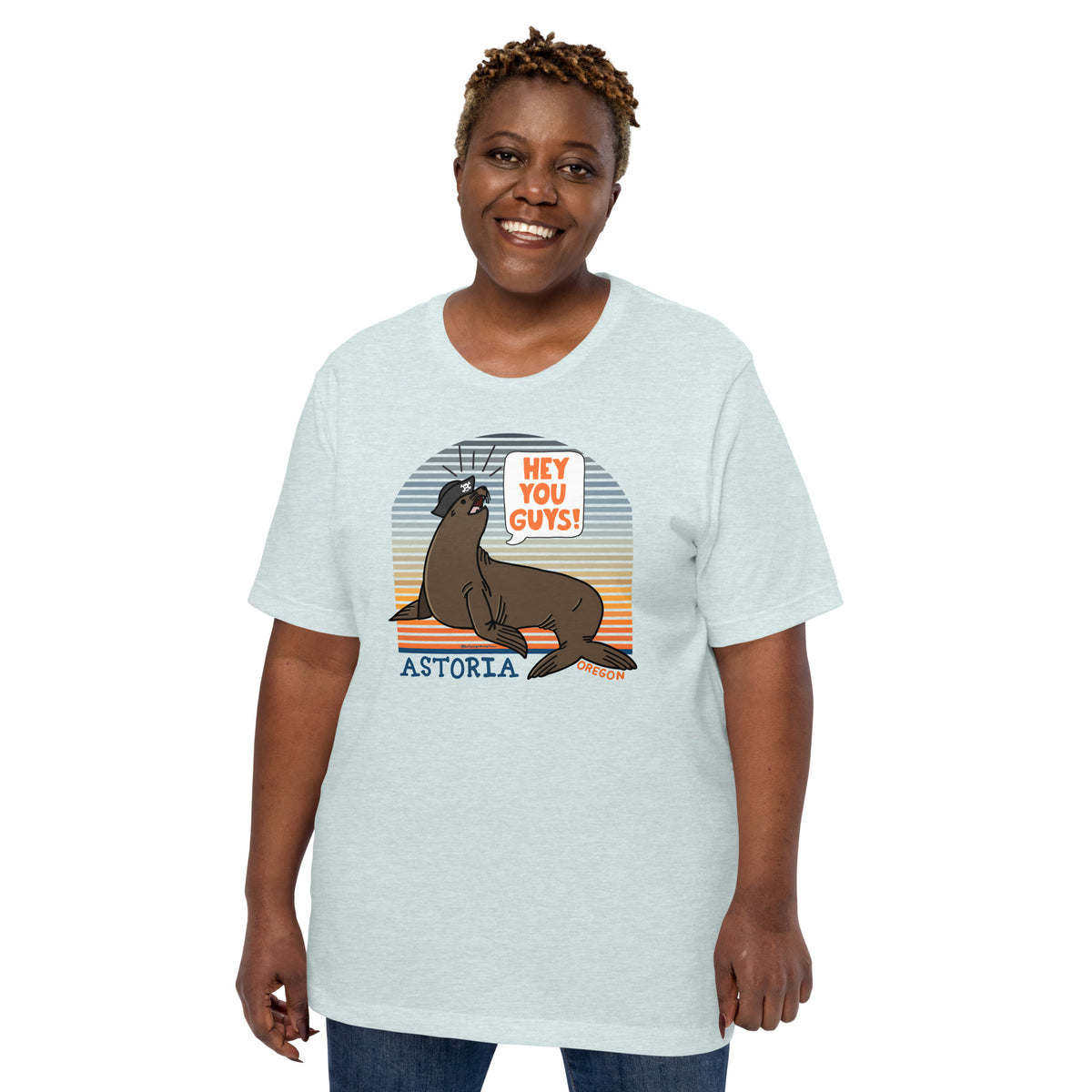A black woman standing in front of a white background wearing jeans and an ice blue t-shirt of a sea lion in a tricorn hat yelling HEY YOU GUYS with a sunset stripe background and hand-lettered Astoria, Oregon on the bottom