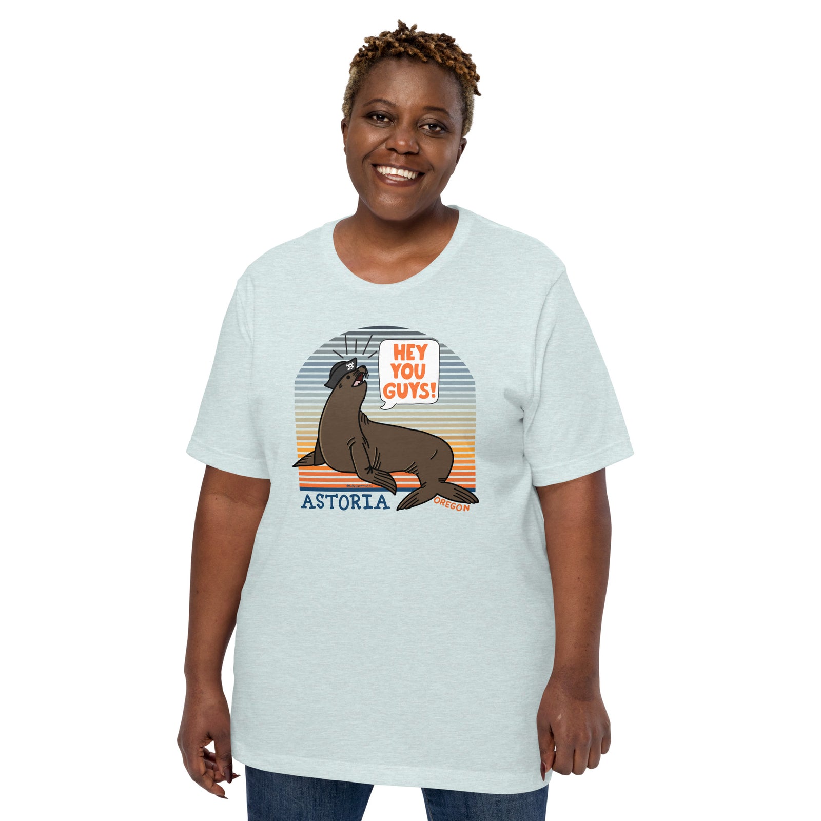 A black woman standing in front of a white background wearing jeans and an ice blue t-shirt of a sea lion in a tricorn hat yelling HEY YOU GUYS with a sunset stripe background and hand-lettered Astoria, Oregon on the bottom
