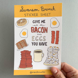 Hand holding Swanson Brunch sticker sheet on a grey background. Sticker sheet has a yellow top and bottom border and has kiss cut stickers of breakfast food, including a sticker with hand-lettered phrase "Give me all the bacon and eggs you have."