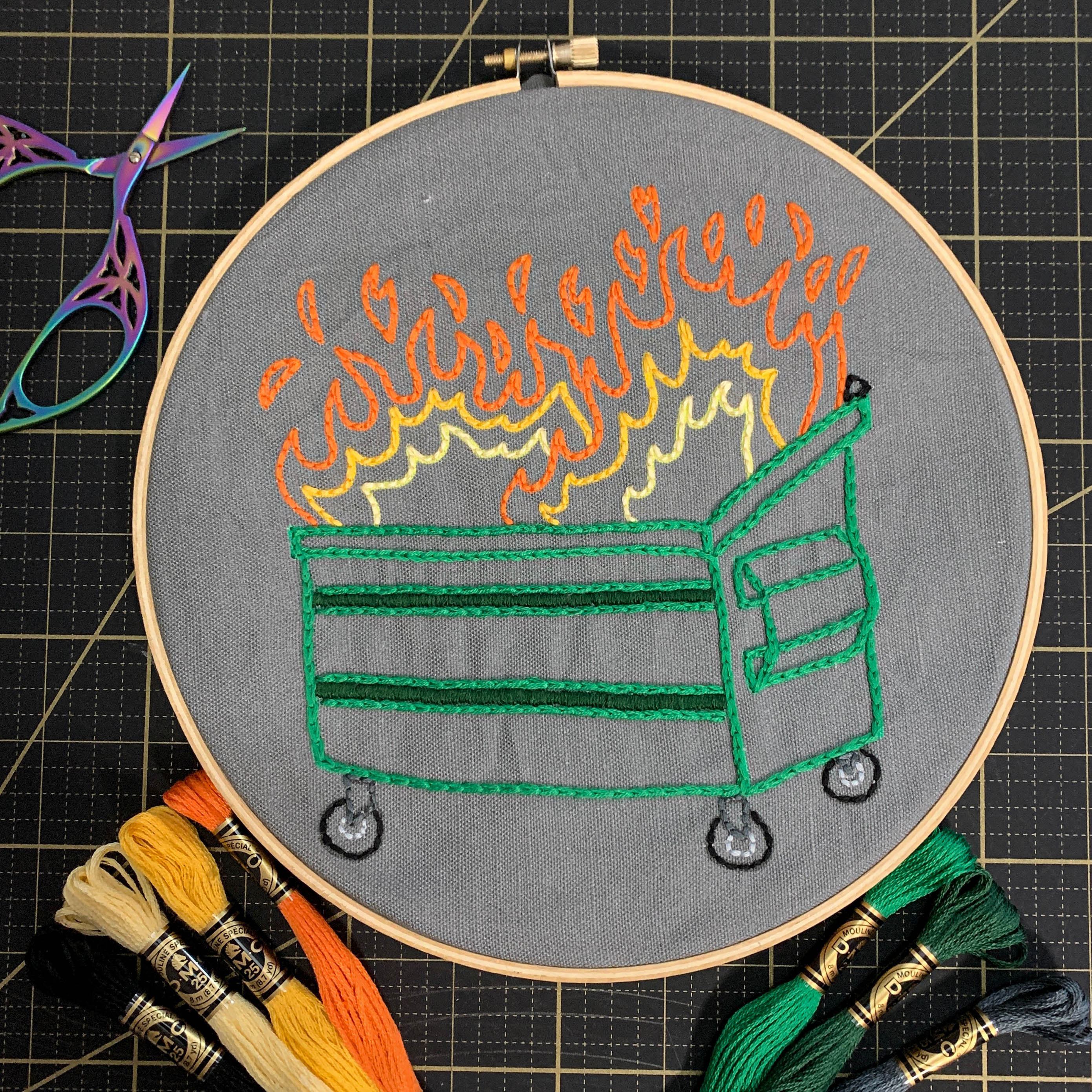 A finished Dumpster Fire embroidery kit in a hoop lays on a black cutting mat. A pair of rainbow metallic embroidery scissors sits open in the top left corner. Each color of embroidery floss is fanned out along the bottom of the embroidery hoop.