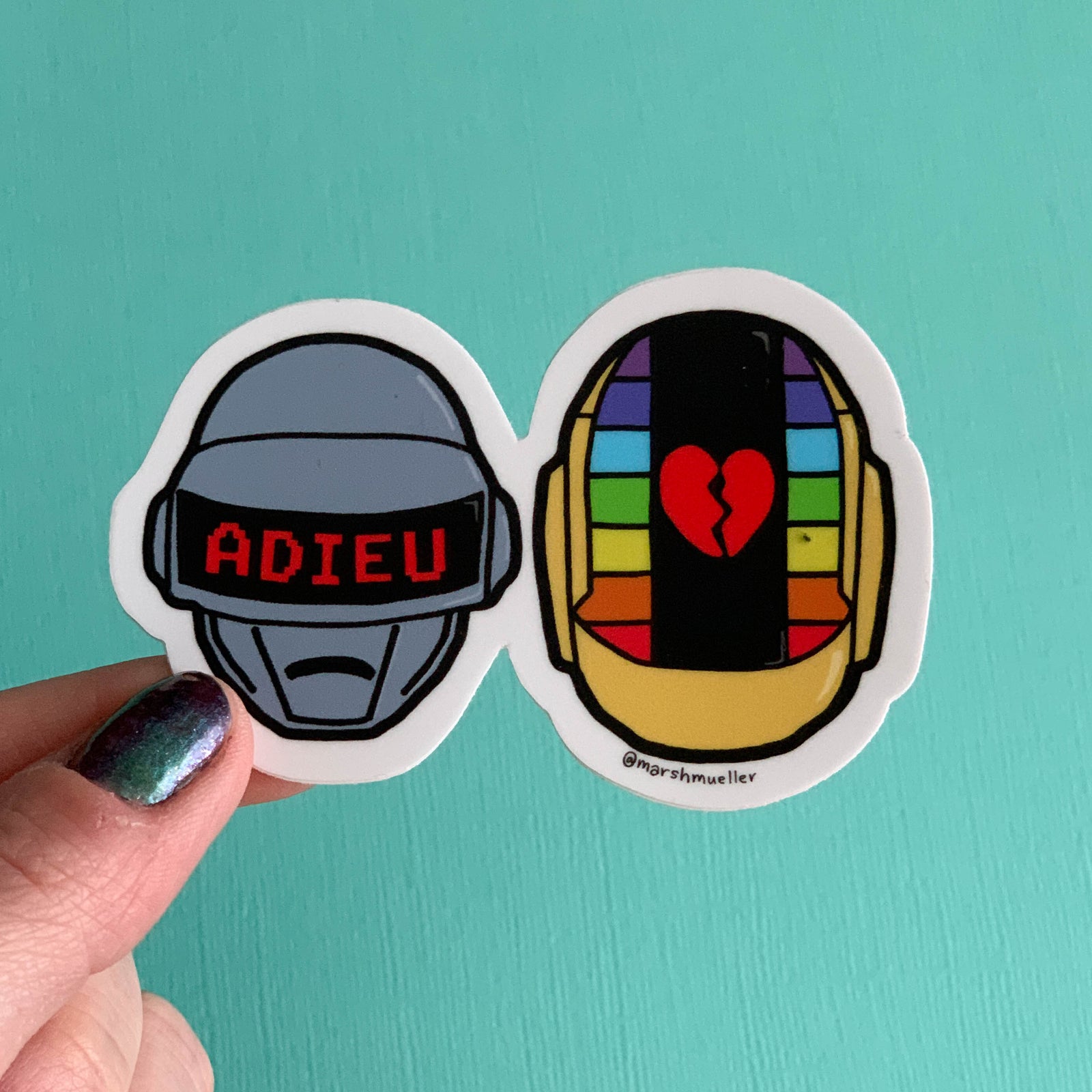 Hand holding a Daft Punk helmets sticker on a teal background
