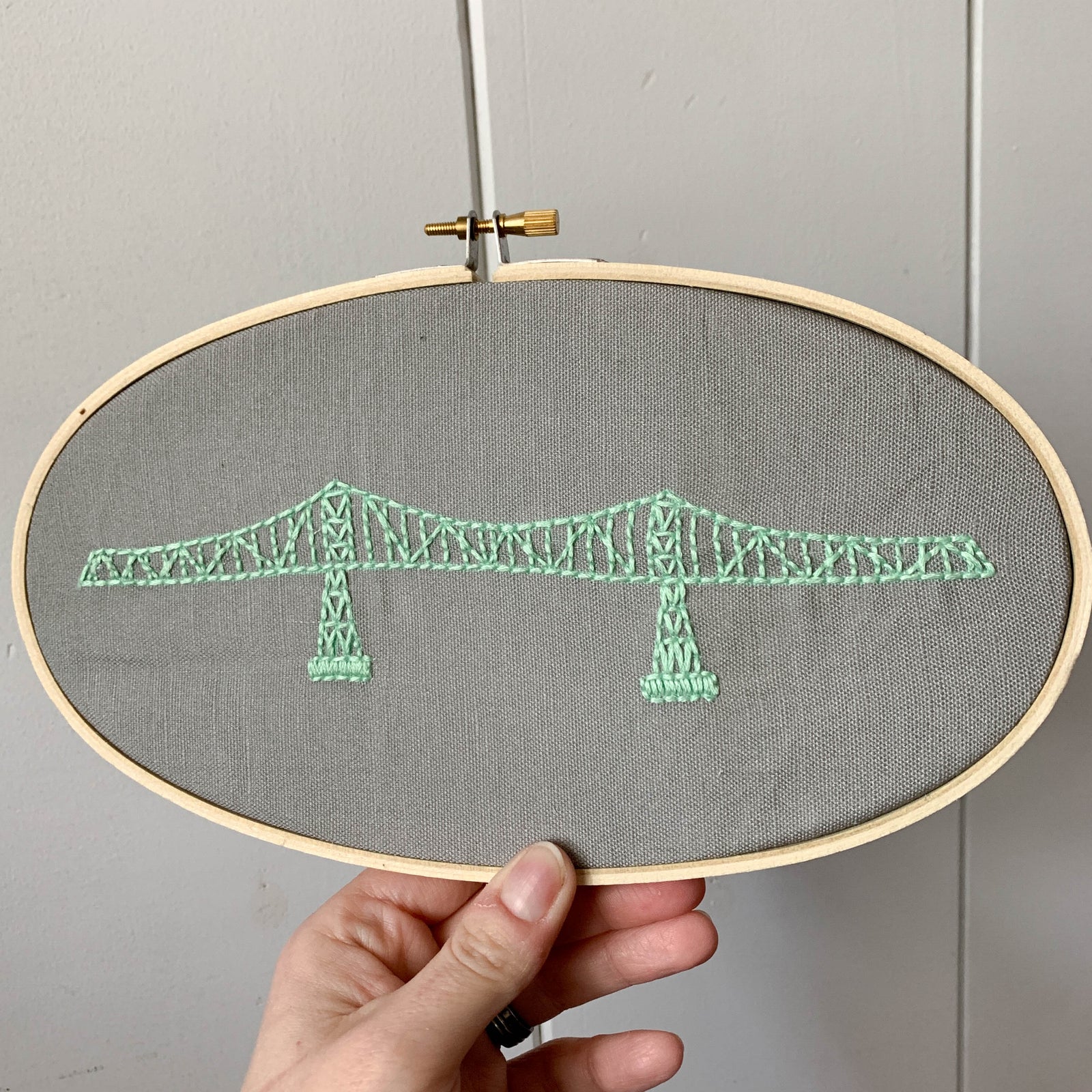 A hand holding a completed Megler Bridge Embroidery Kit with a white wall as the background
